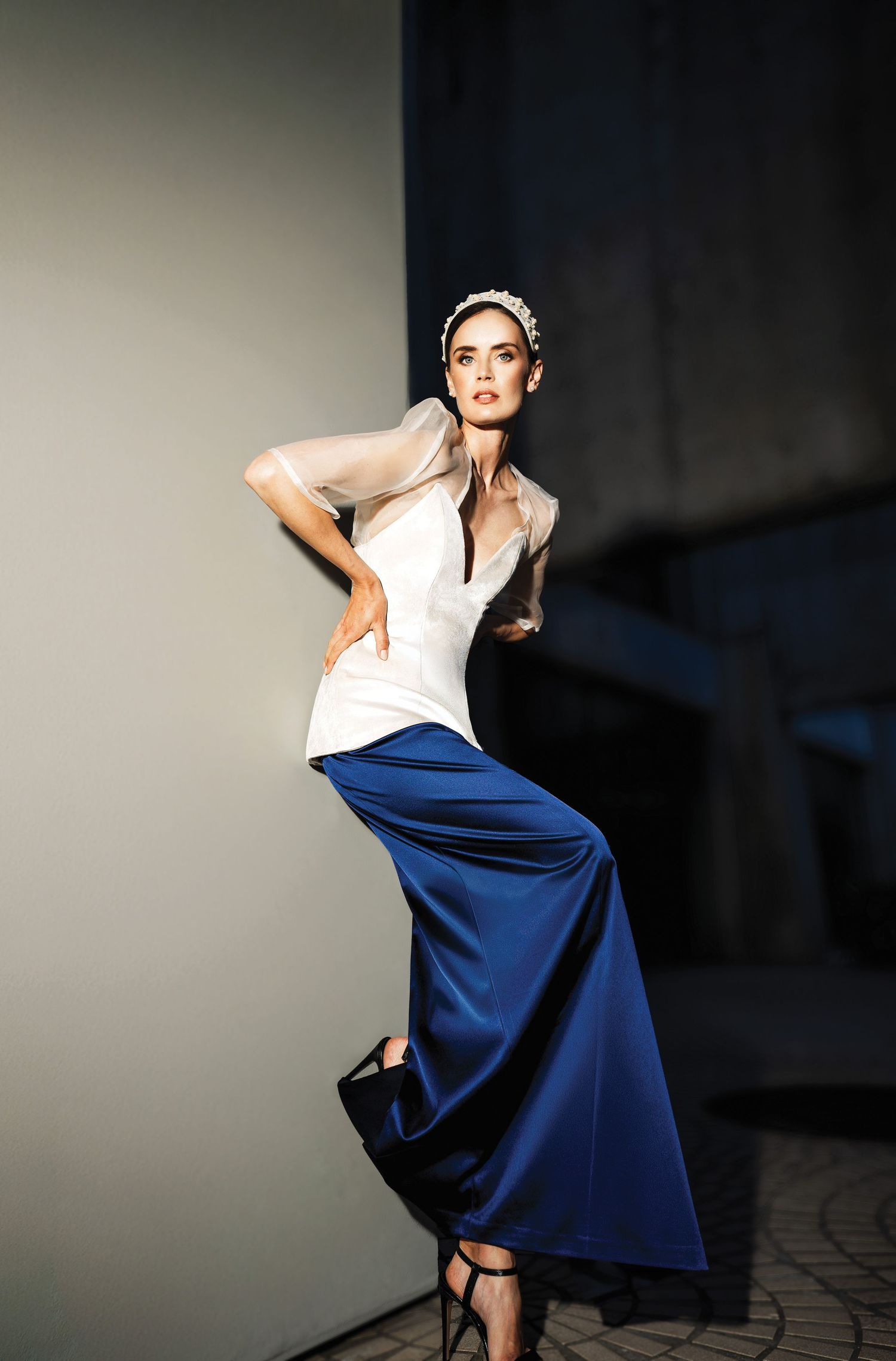 Model poses in white sleeveless top and flowing royal blue skirt with dramatic silhouette against dark background.