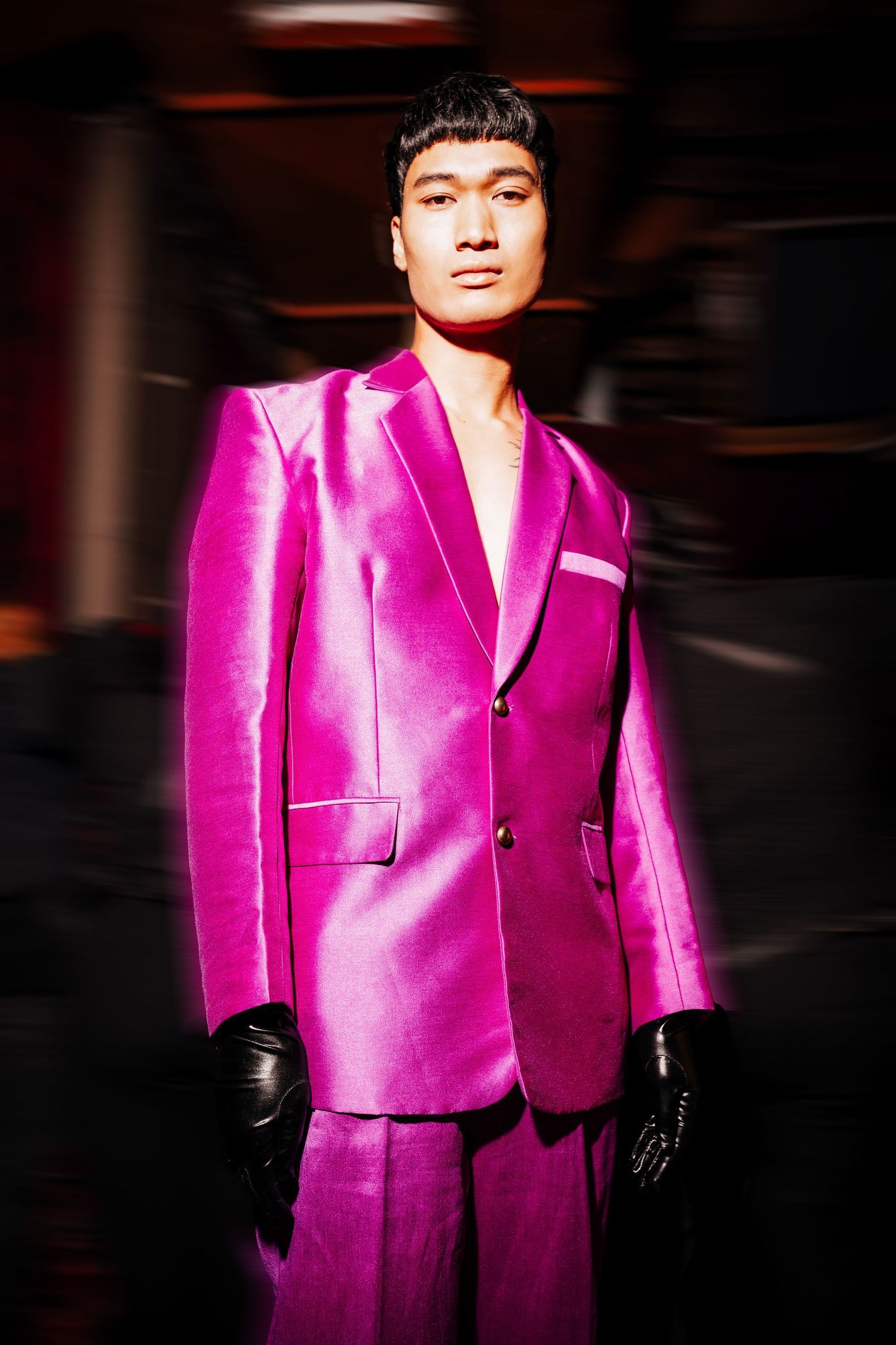 A model walks the runway wearing a vibrant hot pink satin suit with black gloves in a dark setting.