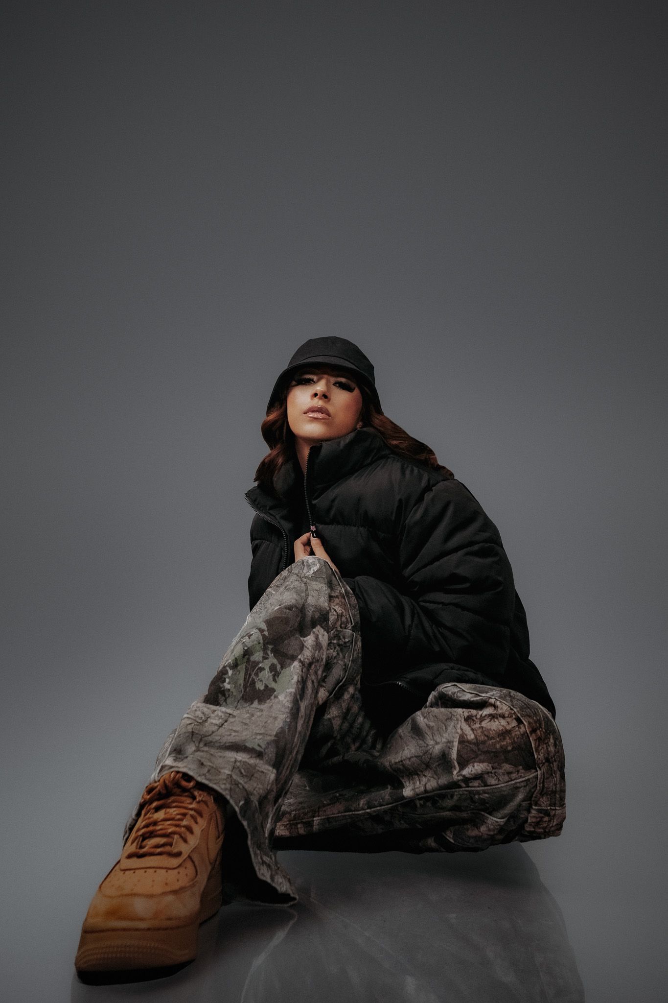 Figure in black puffer jacket and baseball cap poses on reflective surface wearing camouflage pants and brown sneakers.
