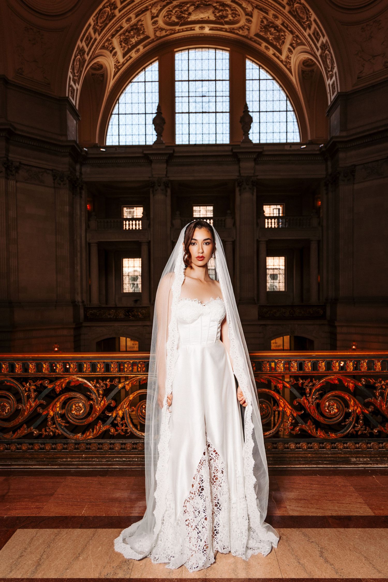 Elegant wedding gown with flowing veil captured in ornate historic building interior with arched window and carved details.