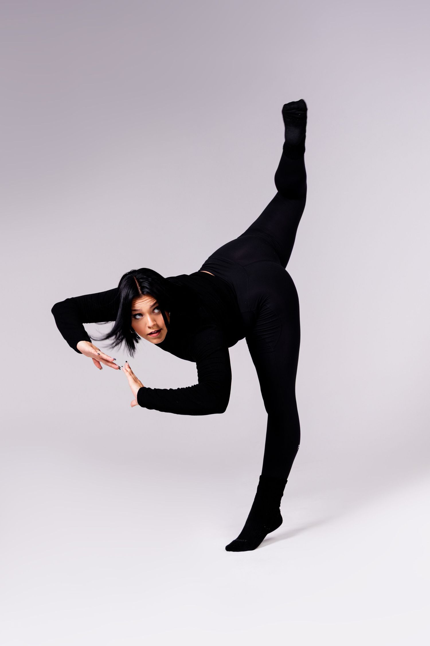 Dancer in black bodysuit performs a high leg extension pose against light studio backdrop.