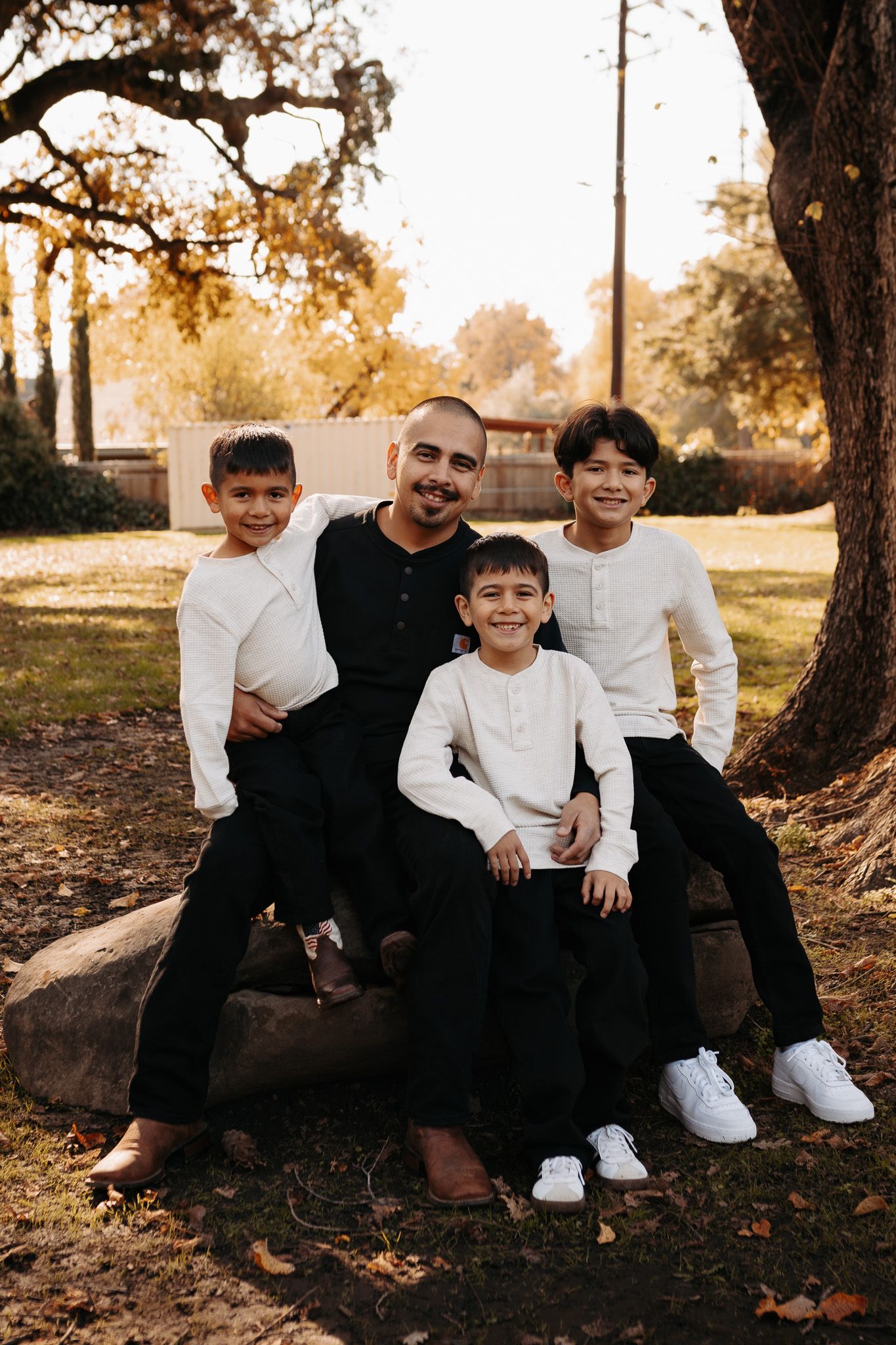 A family group sits together on a wooden bench under an autumn tree at golden hour in a park setting.