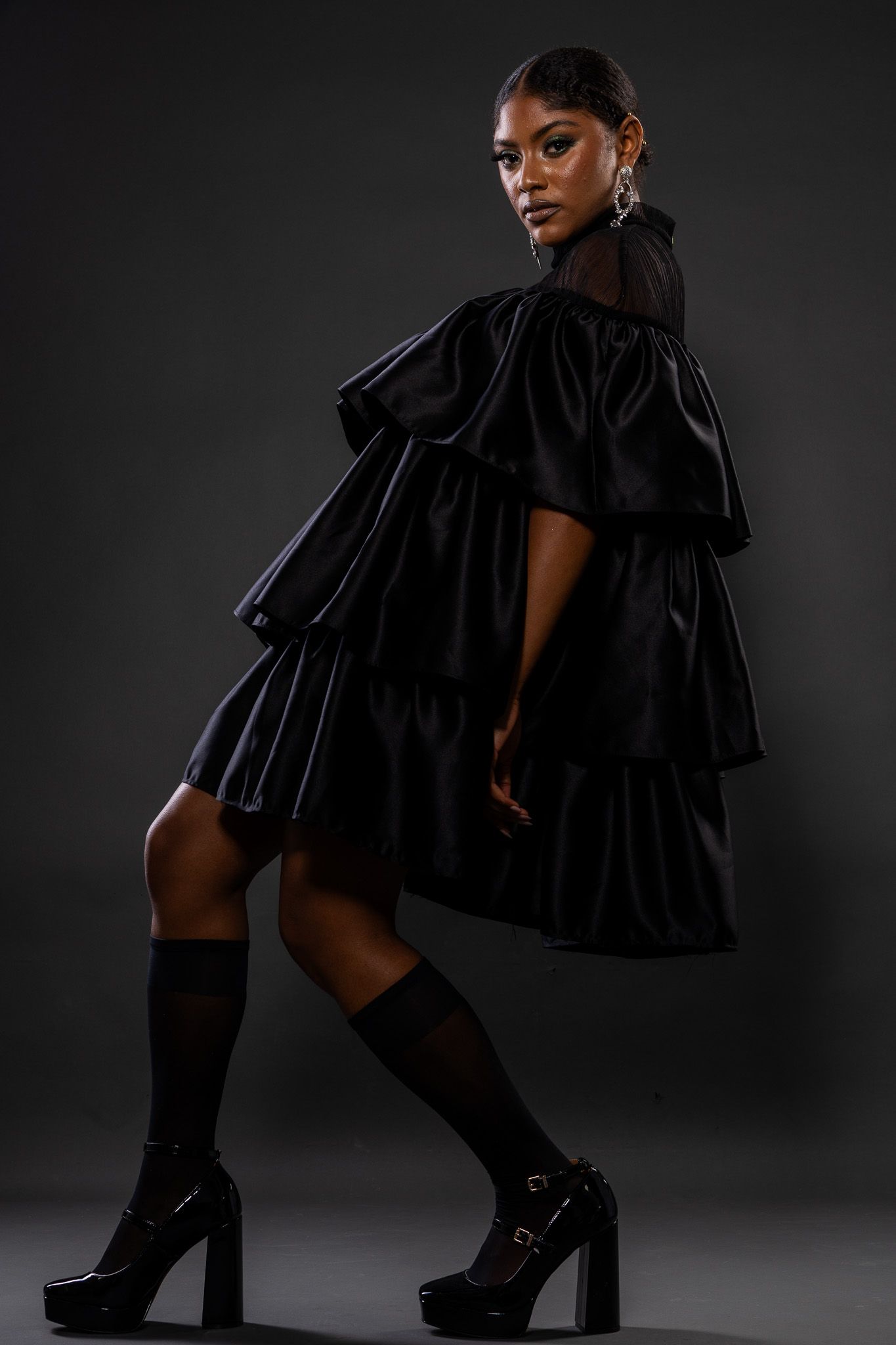 Model wearing a dramatic black tiered ruffle dress with knee-high socks and platform heels against dark background.
