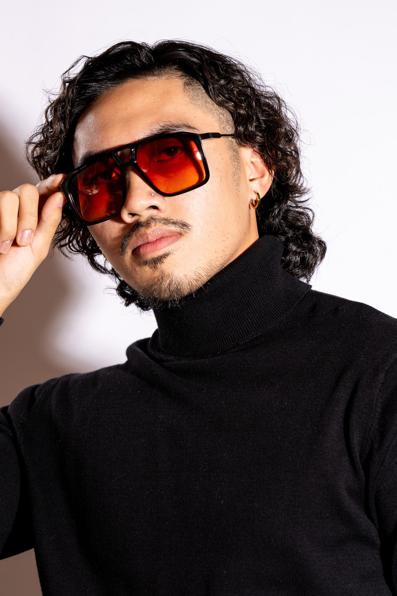 Fashion portrait of a person with curly hair wearing red tinted sunglasses and a black turtleneck sweater.