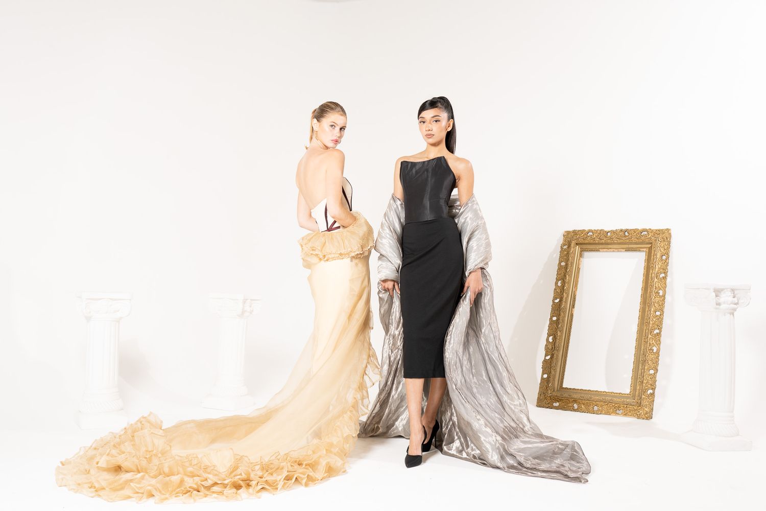 Two people pose in evening gowns with flowing fabrics against white wall with gold mirror frame.