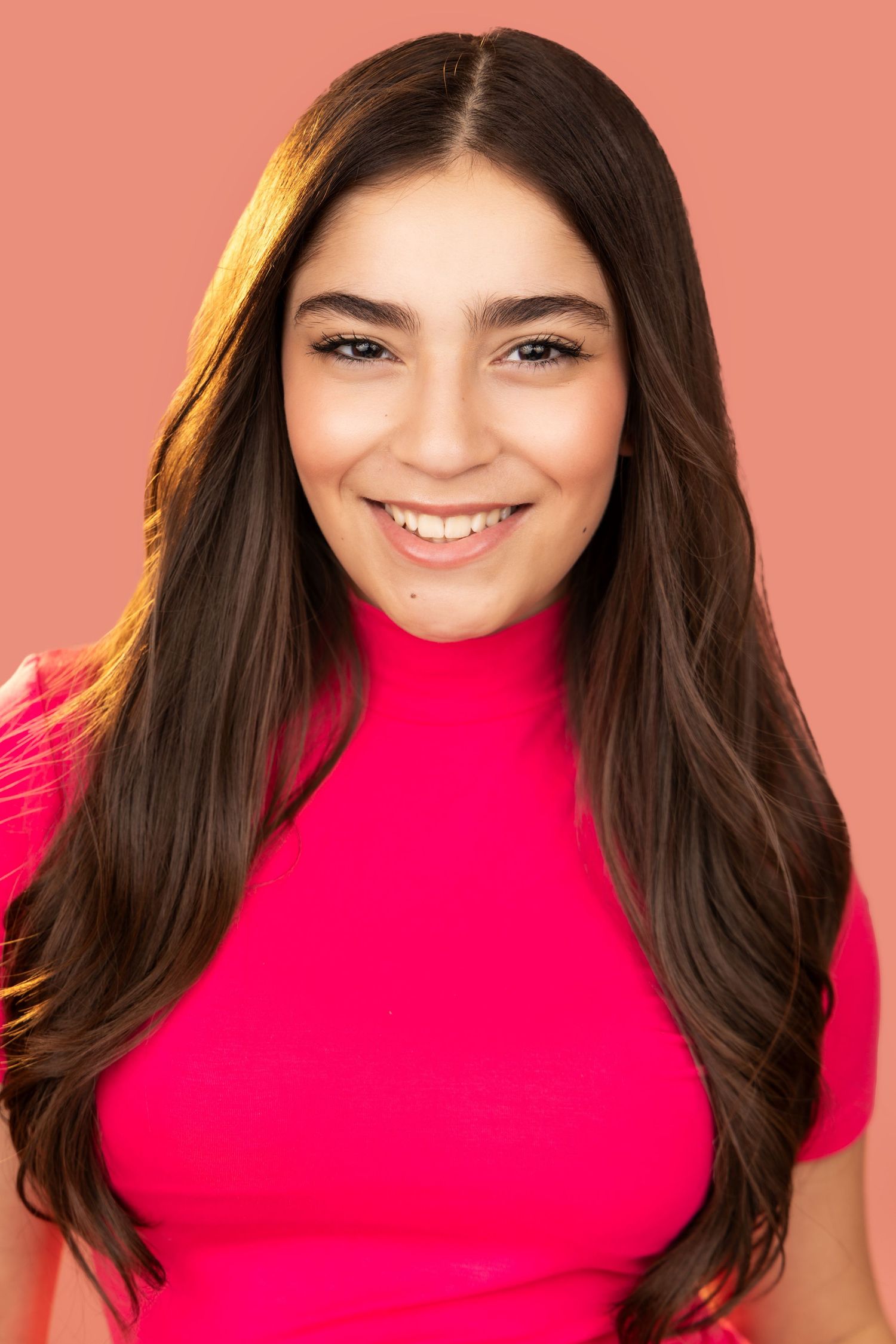 A smiling young woman with long dark hair wearing a bright red turtleneck top against a coral-colored background.