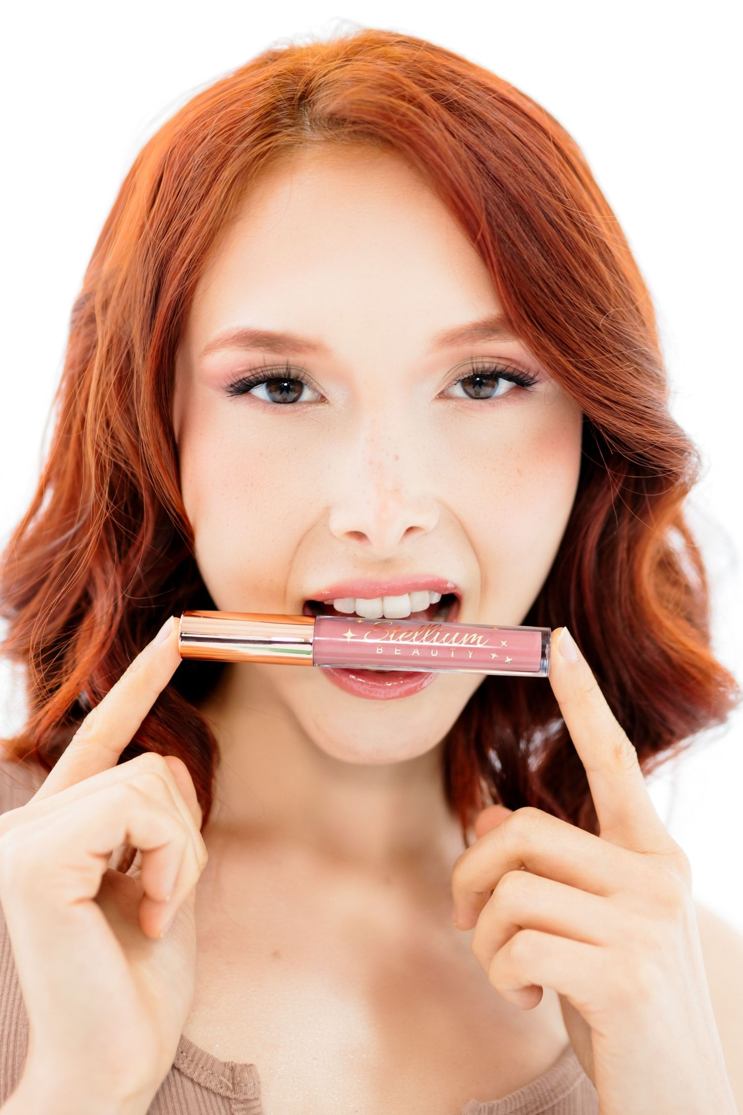 Person with wavy auburn hair holds a makeup product between their teeth while looking at the camera.