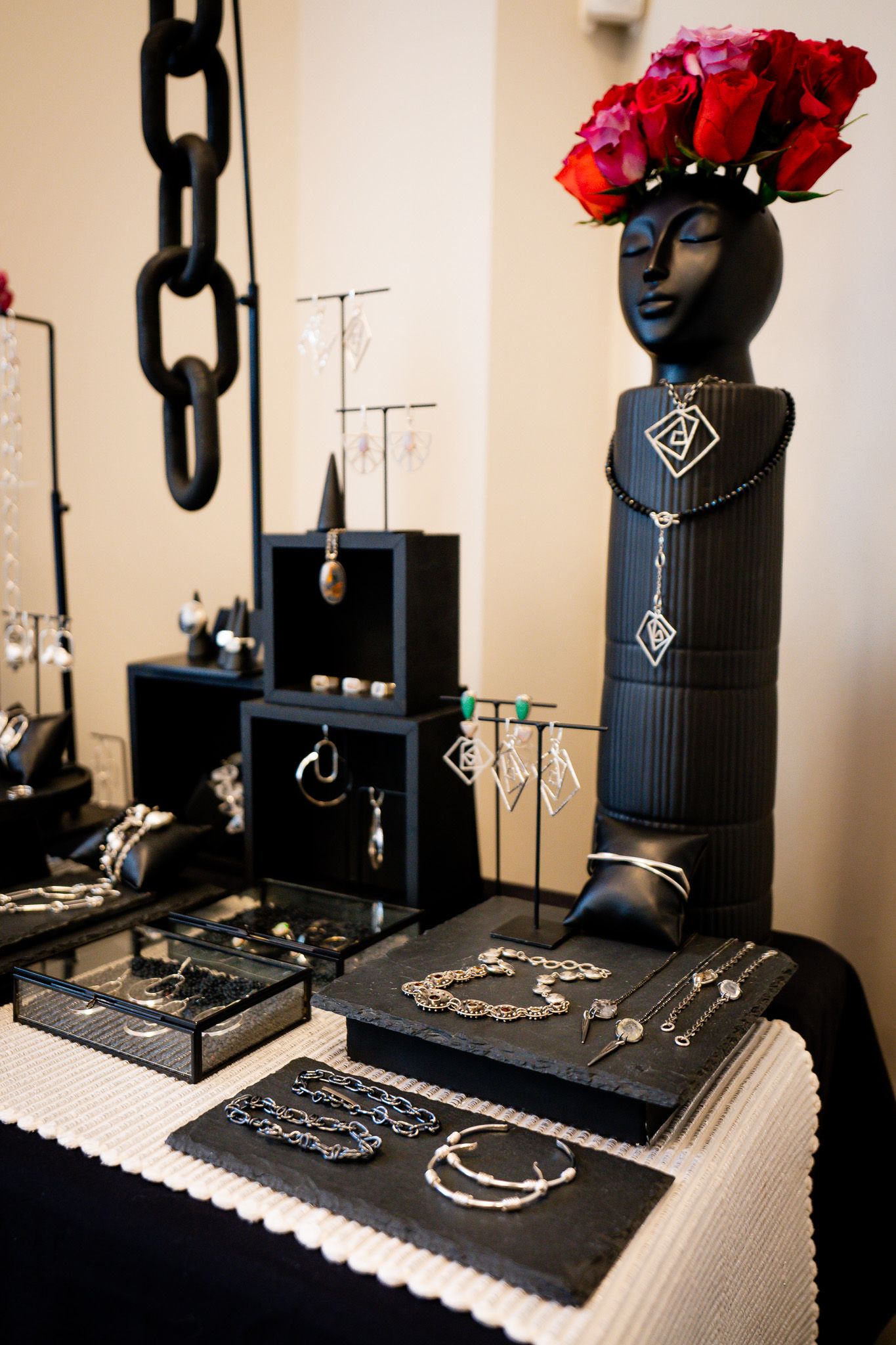 Elegant jewelry display setup with black mannequin bust adorned with red roses and various silver pieces on white tablecloth.