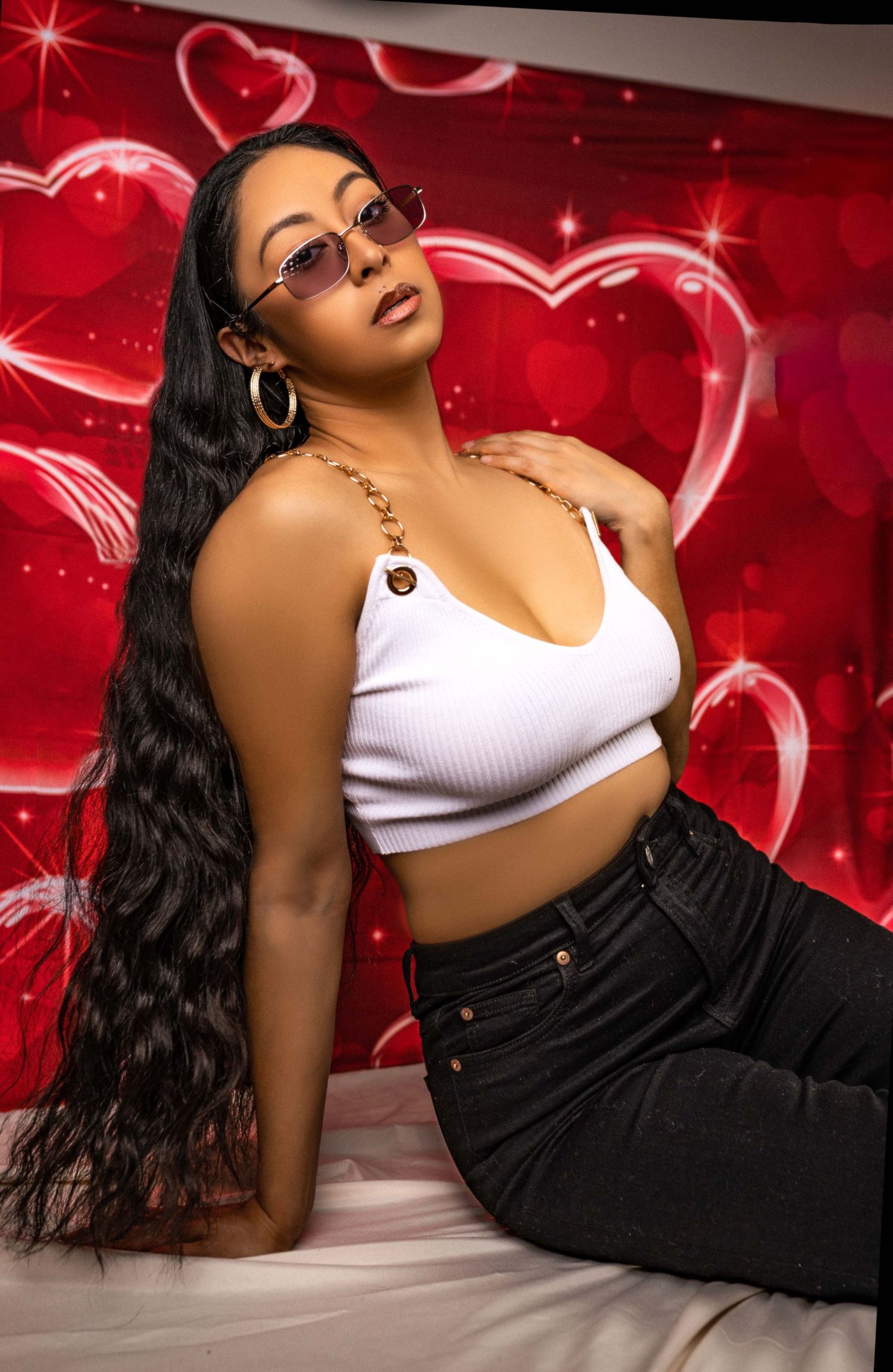 Model posing against red background with glowing heart designs wearing white crop top and dark jeans.