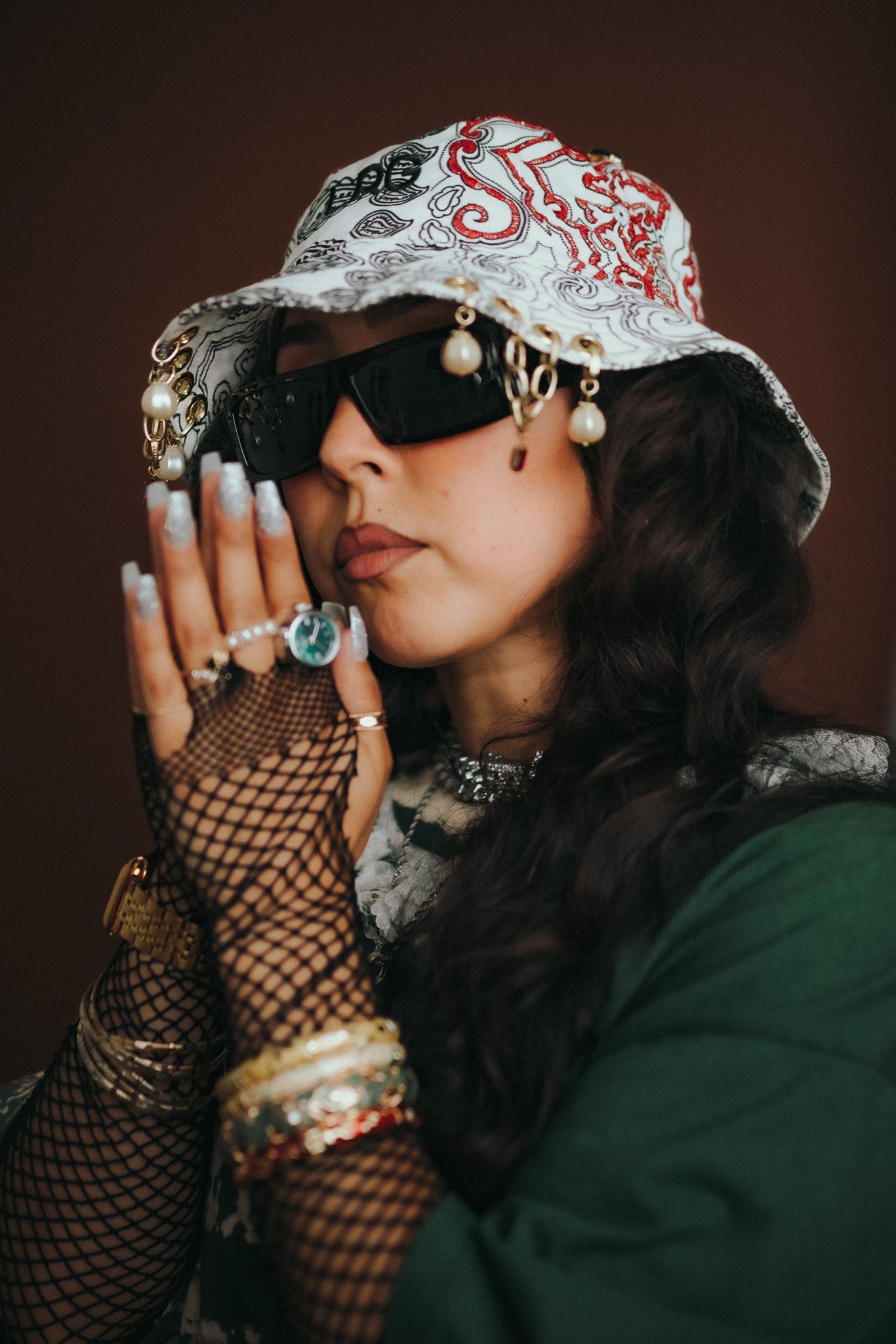 A moody fashion portrait showing fishnet gloves, stacked bracelets, bold bucket hat and dark sunglasses against a dark backdrop.