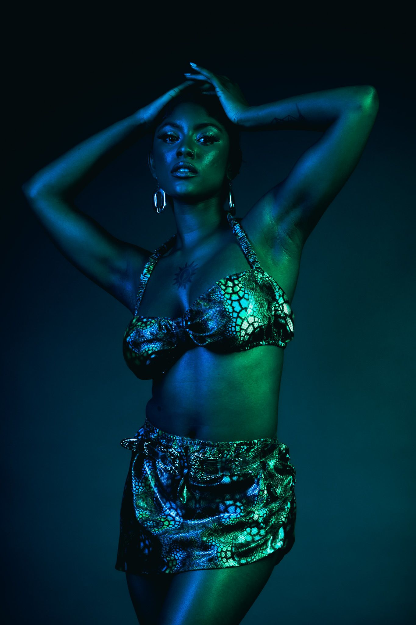 Fashion model poses in metallic patterned bikini under dramatic blue and green theatrical lighting.