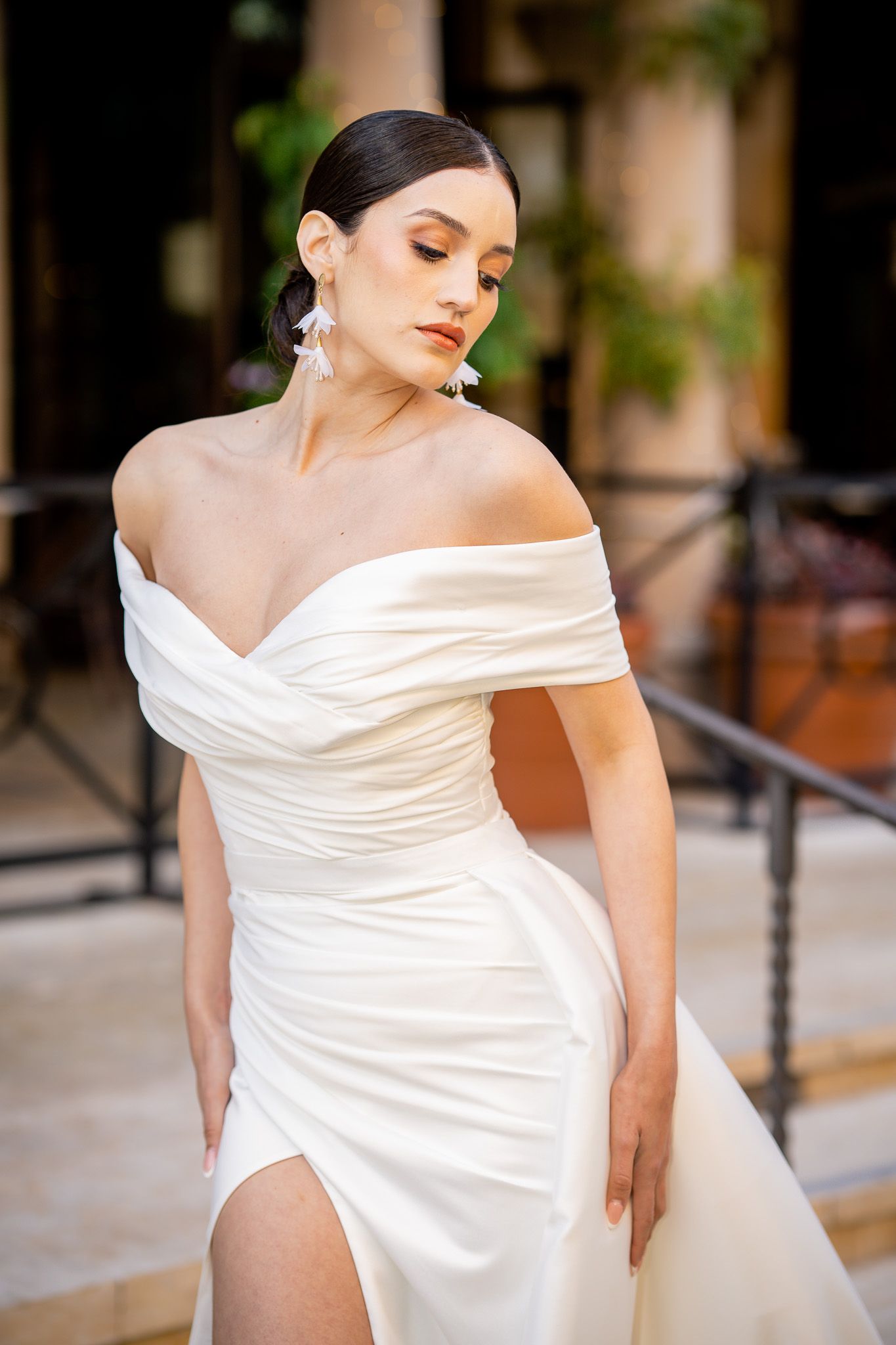 A wedding dress model poses in an elegant off-shoulder white gown with ruched detailing and a high slit.