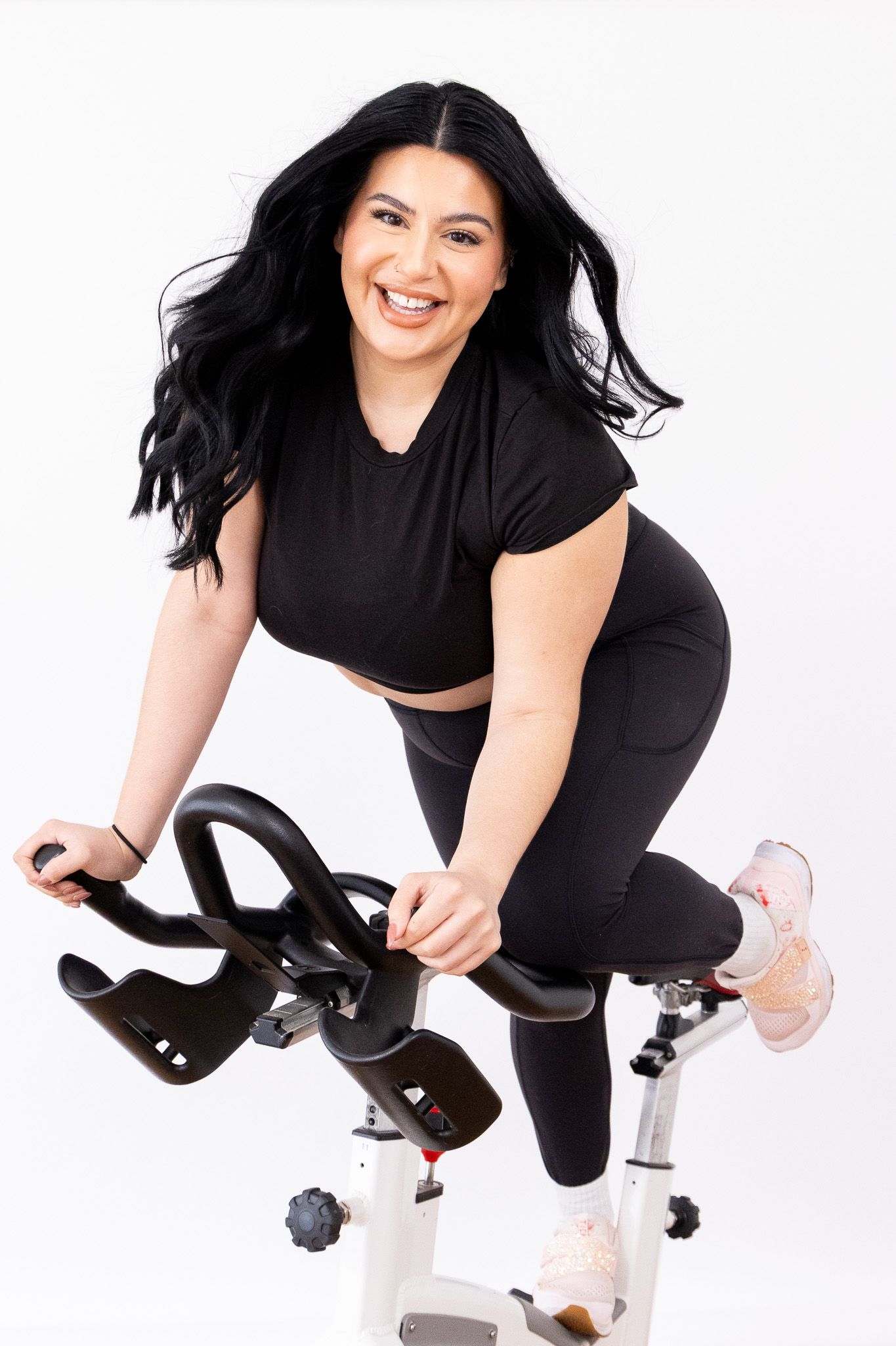 A woman in black athletic wear smiles while exercising on a spin bike against a white background.