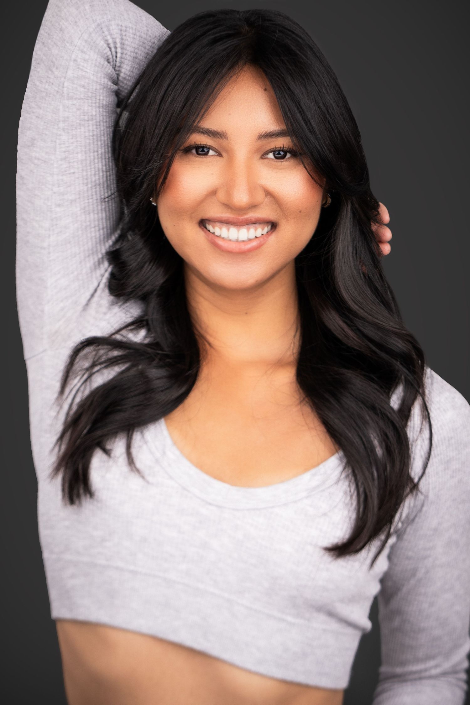 A young Asian woman wearing a grey crop top smiles warmly against a dark background.