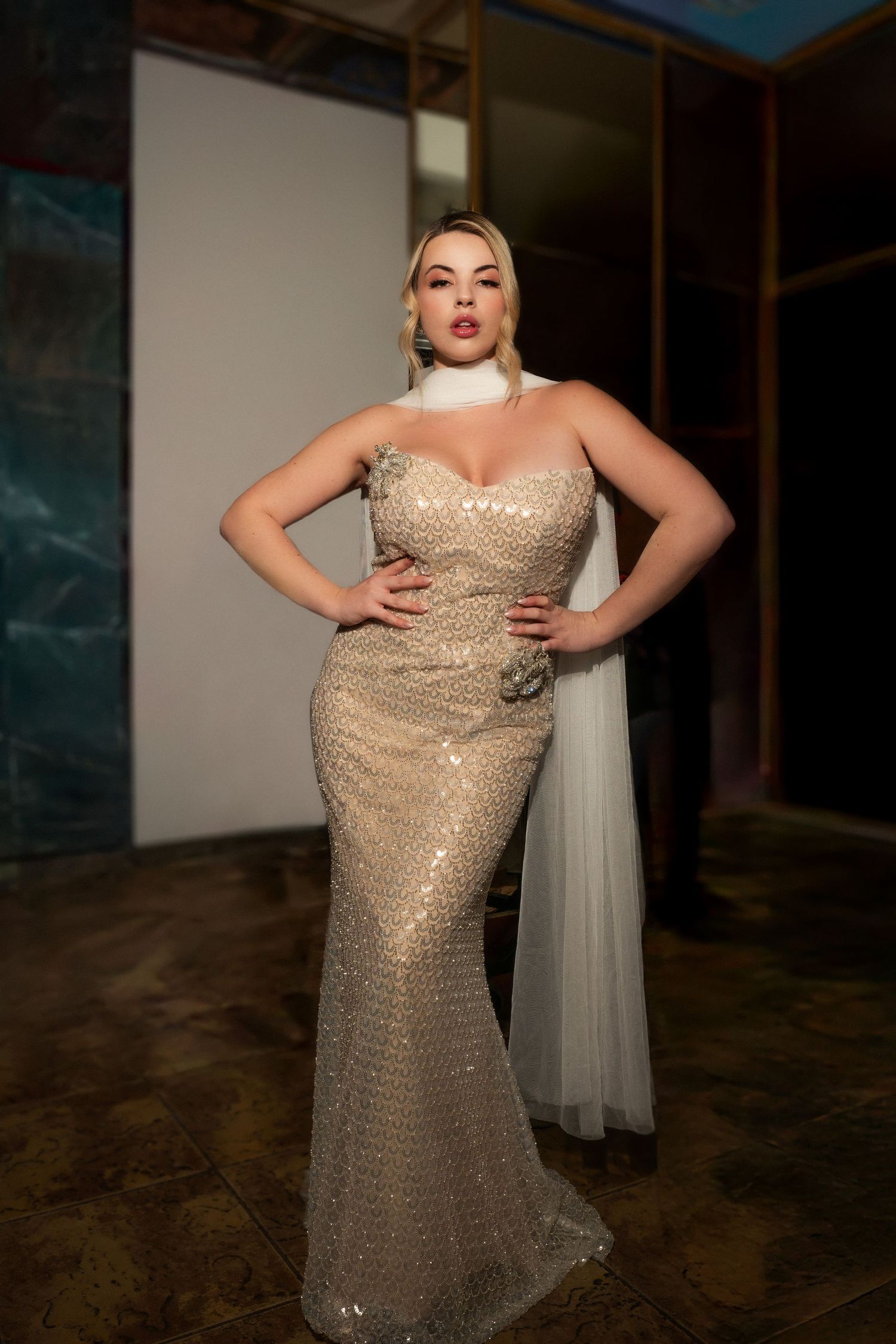 A model poses in a form-fitting champagne sequined evening gown with a sweetheart neckline in a dimly lit room.