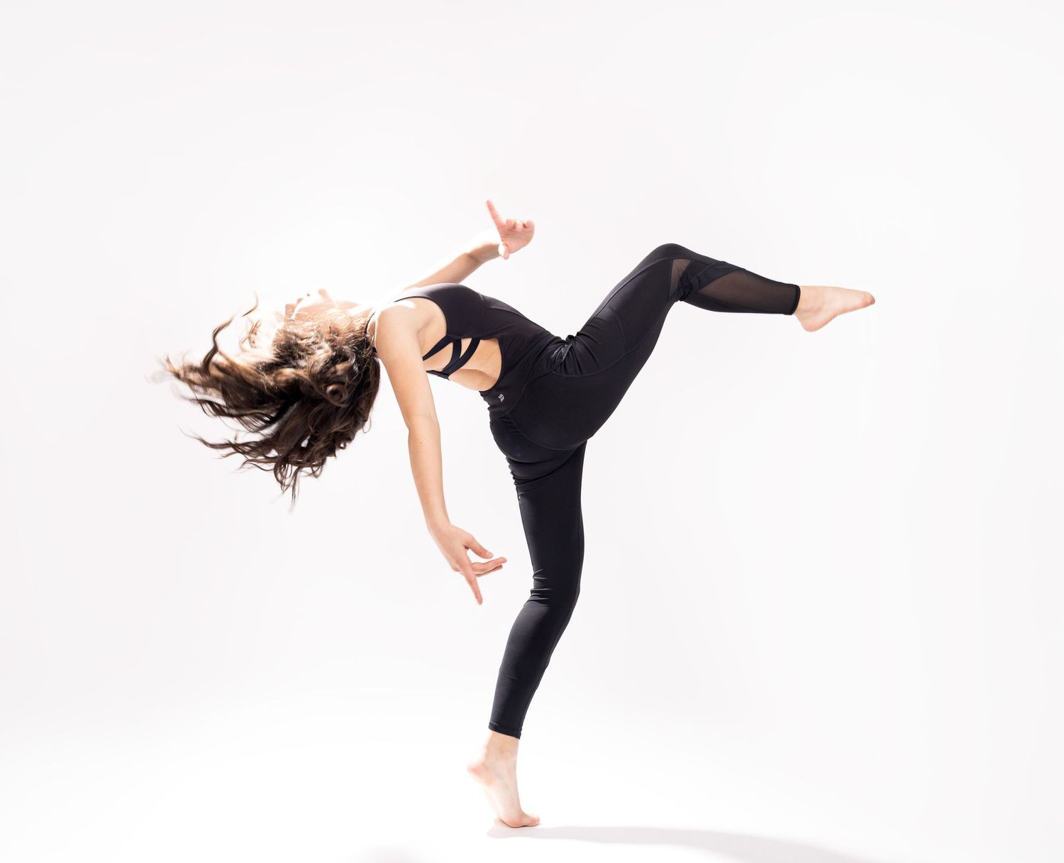 A dancer in a black leotard and leggings performs a dynamic high kick pose against a white background.