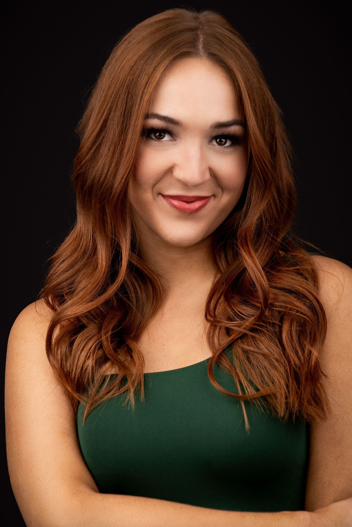 Studio portrait of a warm smile against a dark background wearing an emerald green sleeveless top with long wavy brown hair.
