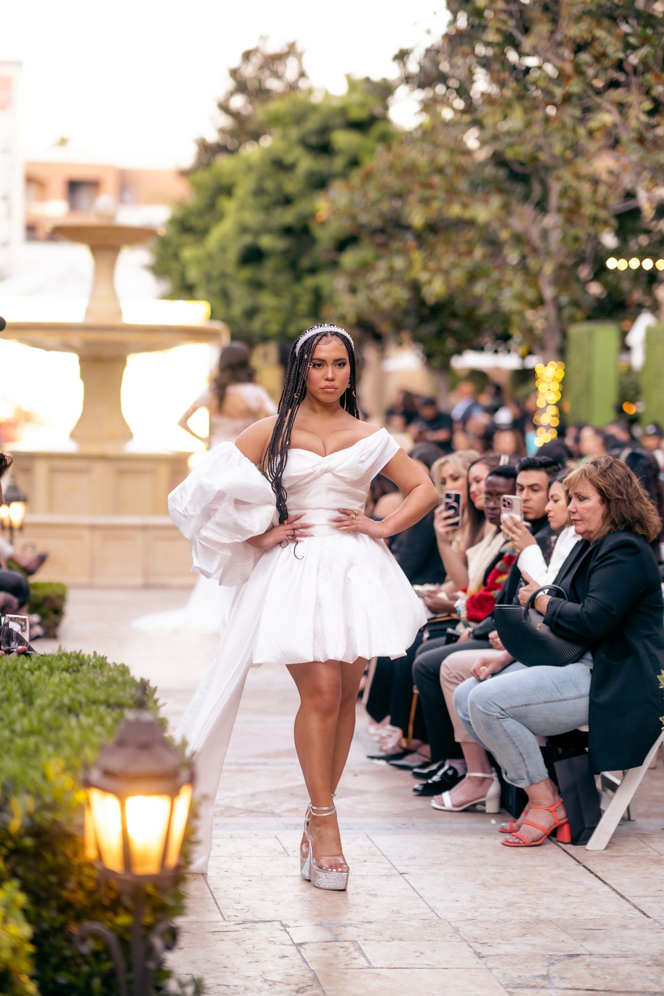 A model walks the runway in a short white cocktail dress with dramatic sleeves during an outdoor evening fashion show.