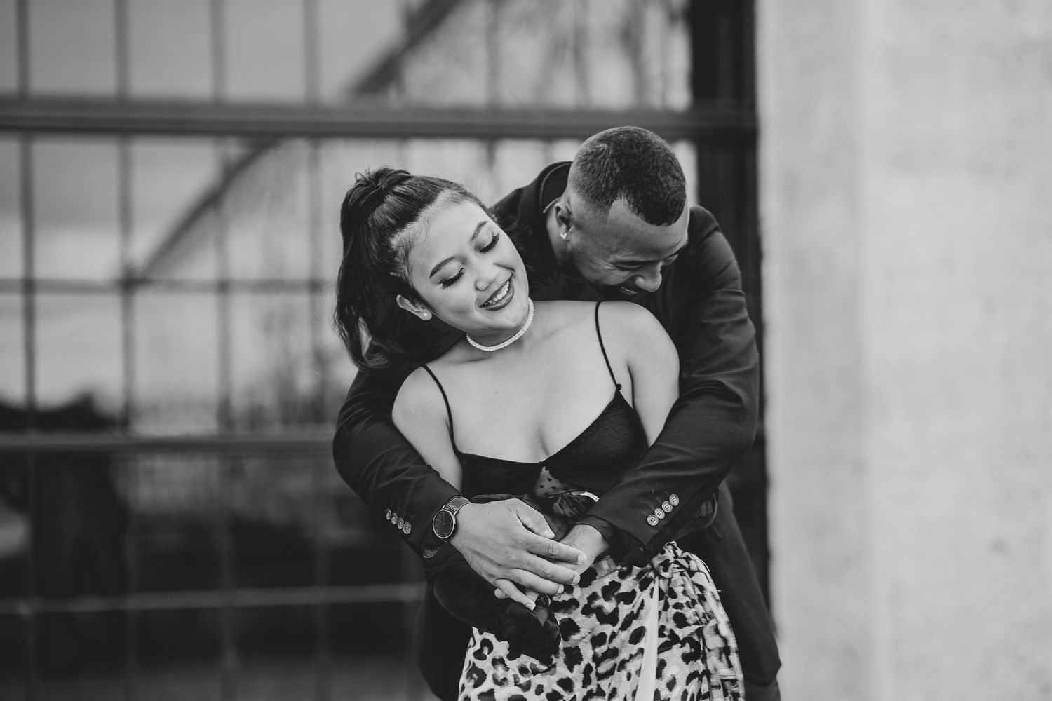 Black and white photo of couple in an intimate embrace, with person wearing a leopard print skirt laughing joyfully outdoors.