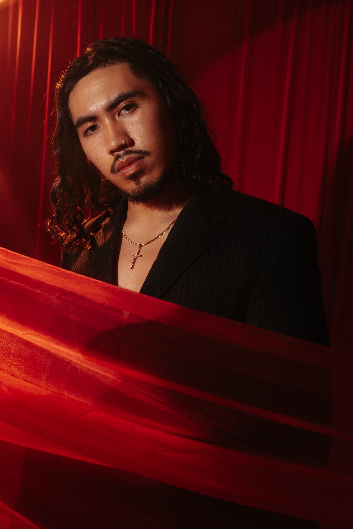 Dramatic portrait with red curtain backdrop showing model with long dark hair in black jacket illuminated by moody lighting.