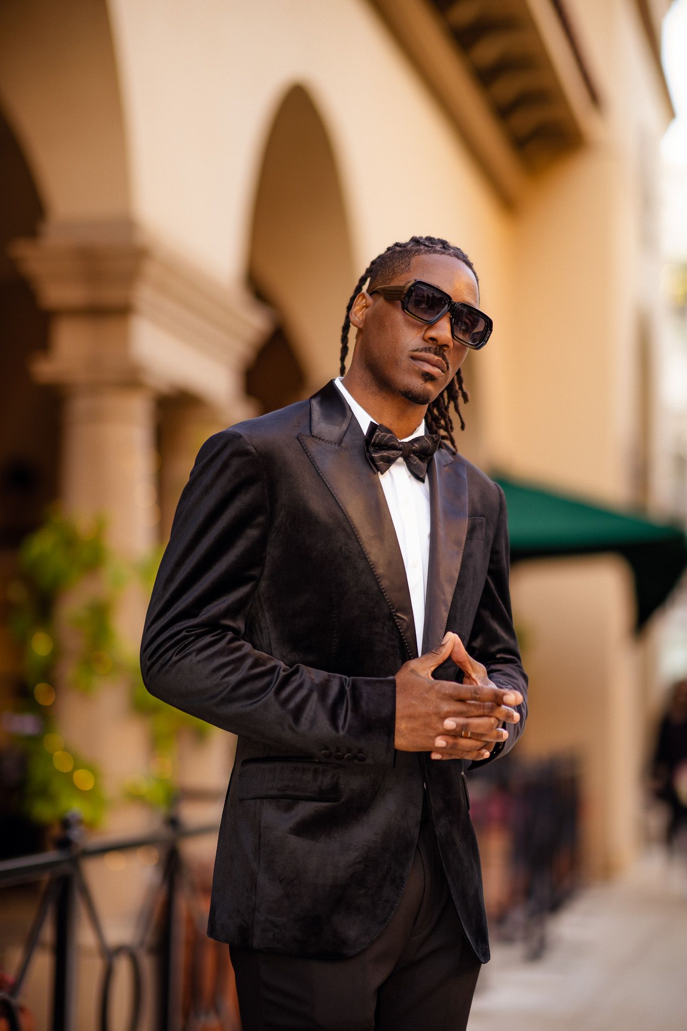 Stylish man in black tuxedo and bow tie poses outdoors wearing sunglasses against architectural backdrop.