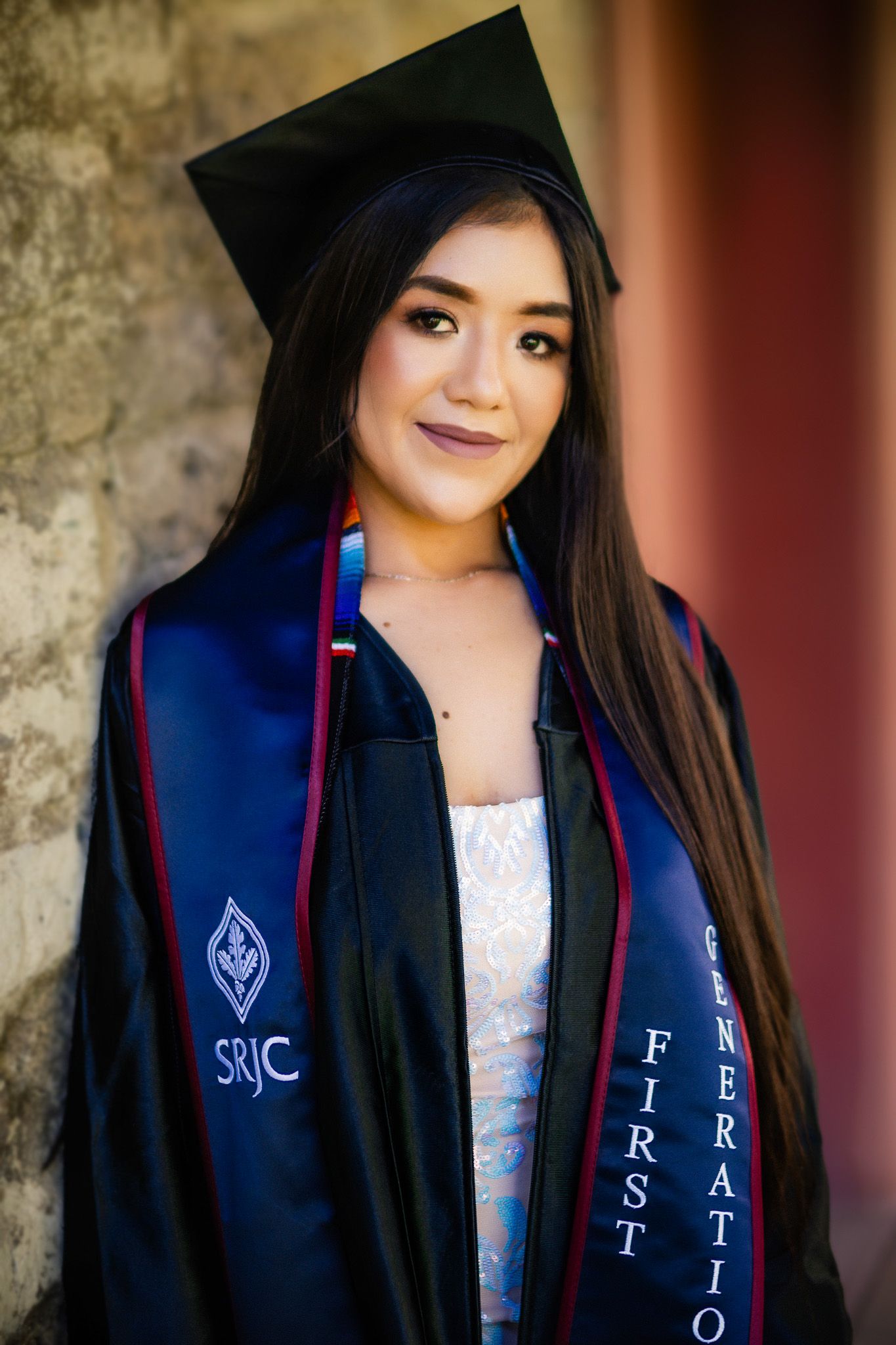 A graduate wearing black cap and gown with SJMC First Generation stole stands against a textured wall for a portrait.