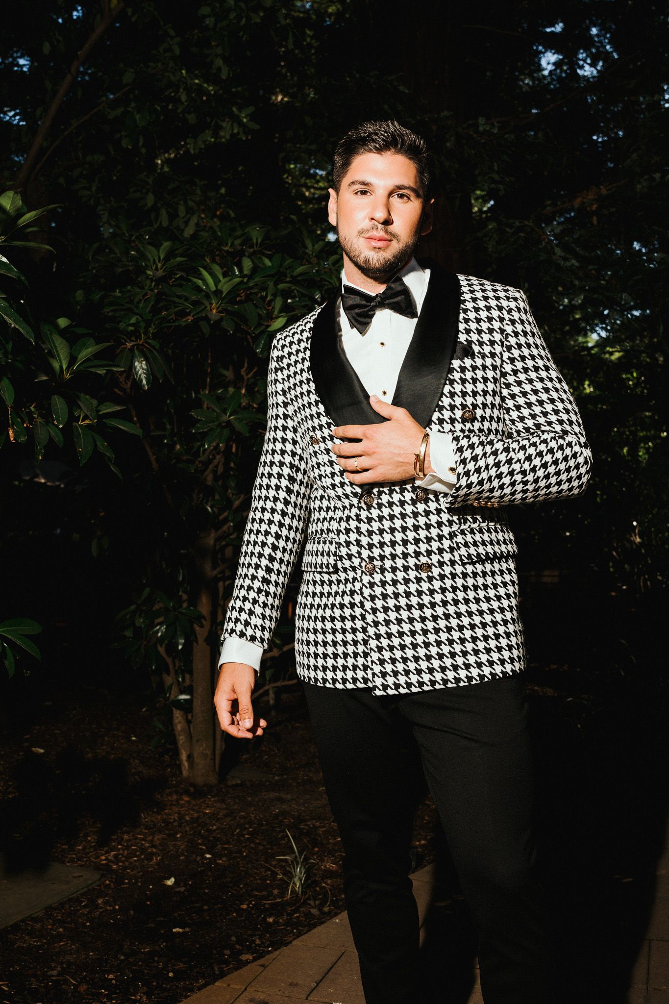 A person wearing a black and white houndstooth suit jacket with bow tie poses elegantly in an outdoor evening setting.
