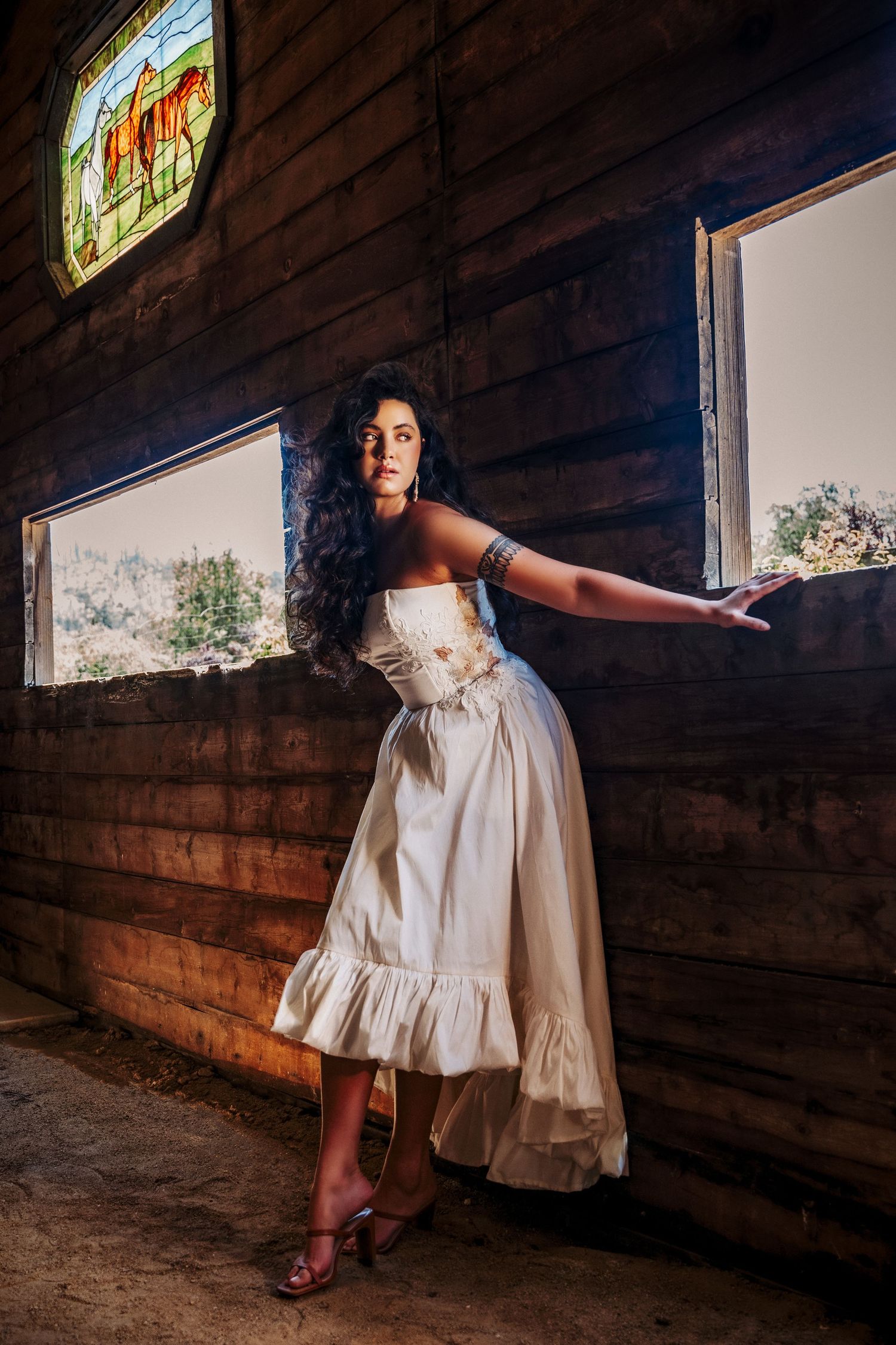 A model in a vintage-style cream dress strikes a graceful pose inside a rustic wooden barn with stained glass windows.