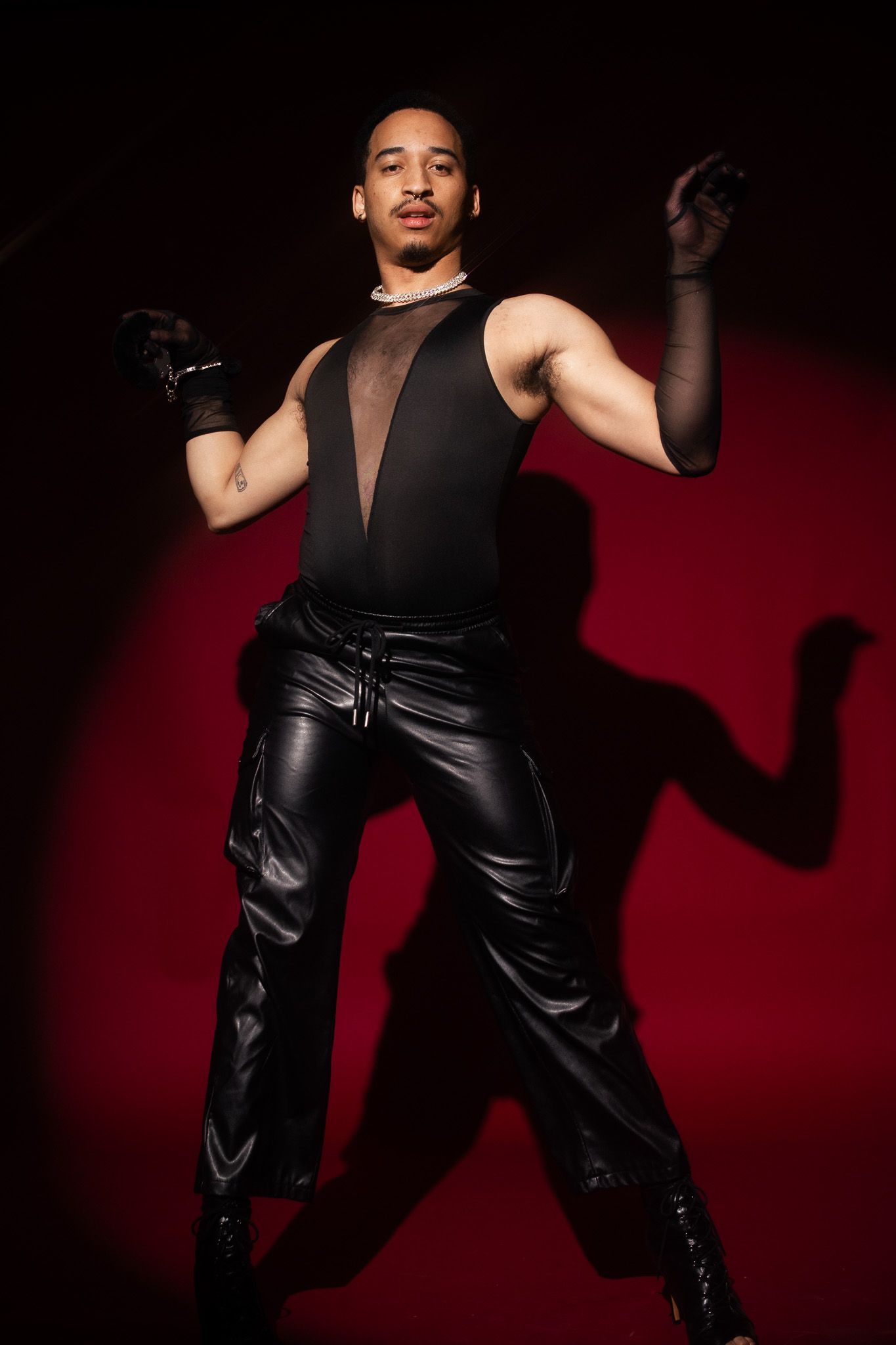 Performer in black sleeveless top and leather pants strikes dramatic pose against dark red background.