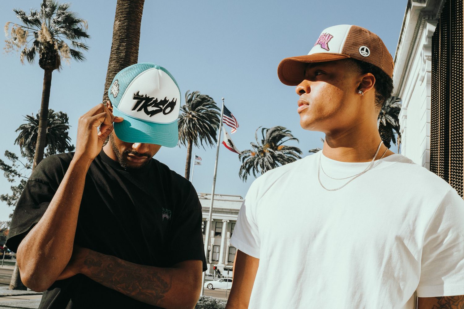 Two people in casual wear standing outdoors with palm trees, one wearing a Nema hat and one in a brown cap.
