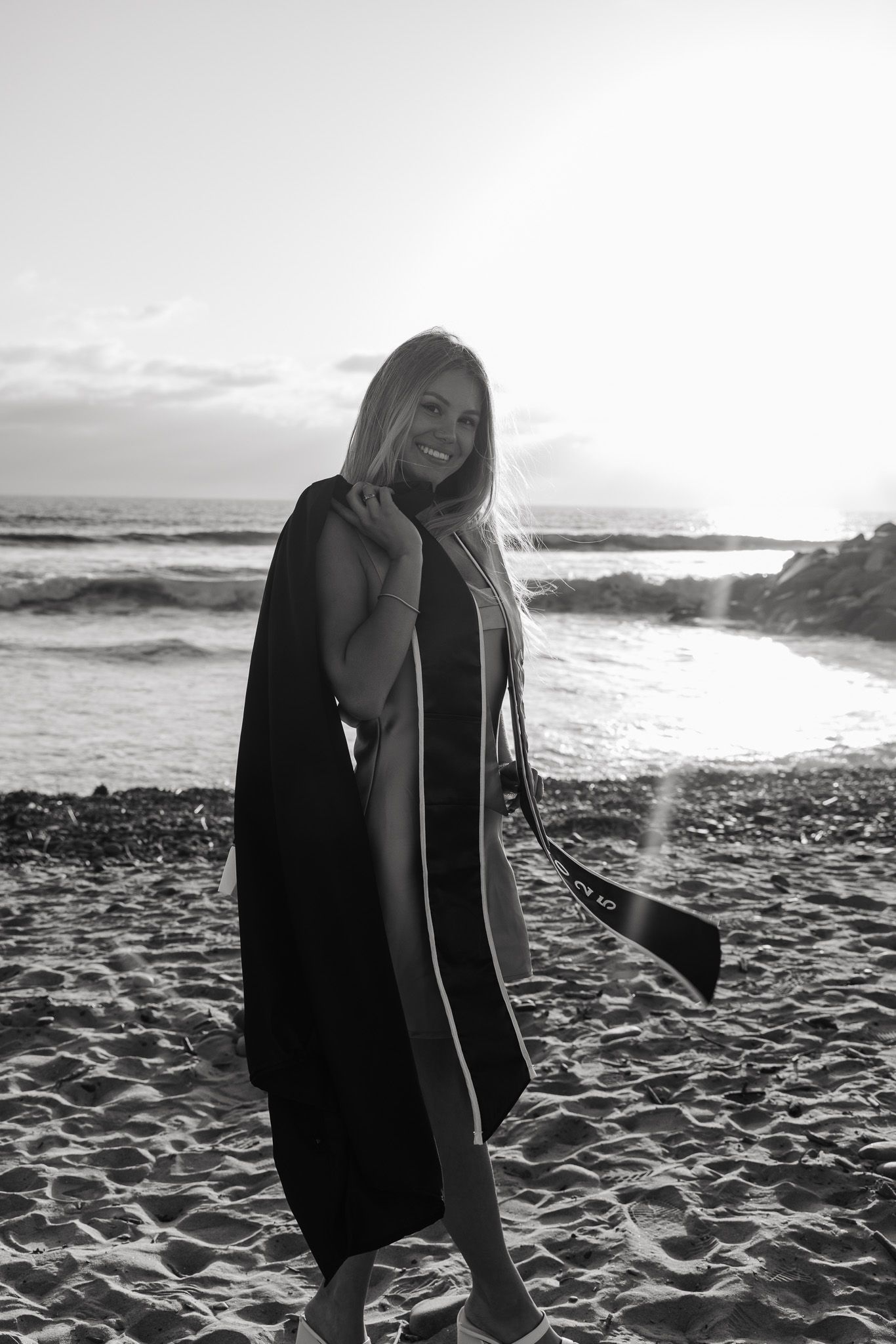Black and white beach photo of someone wearing a long dark robe standing on sand with ocean waves in background at sunset.