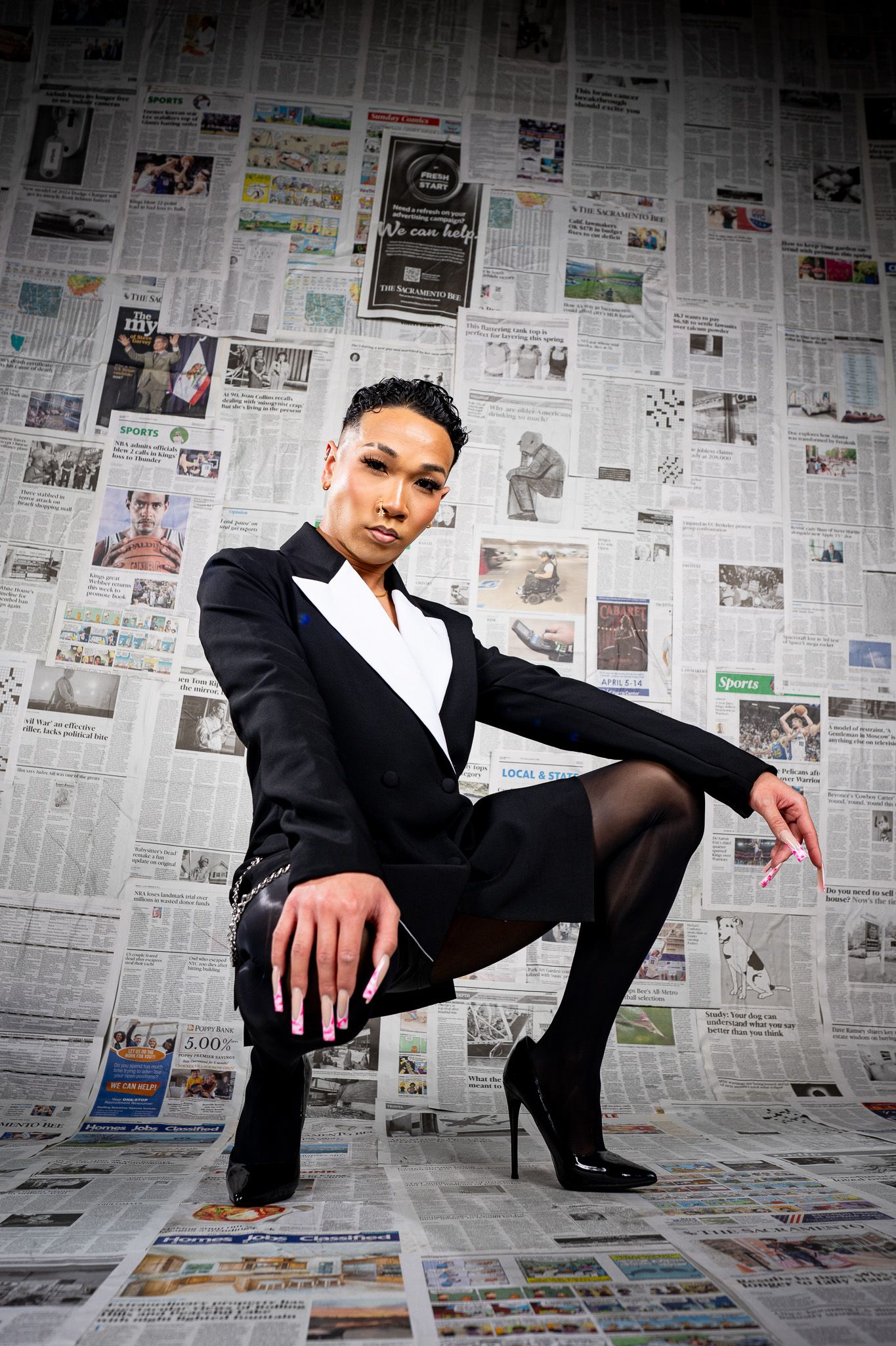 Model in black suit ensemble and heels poses against a wall of newspaper clippings in dramatic editorial shoot.