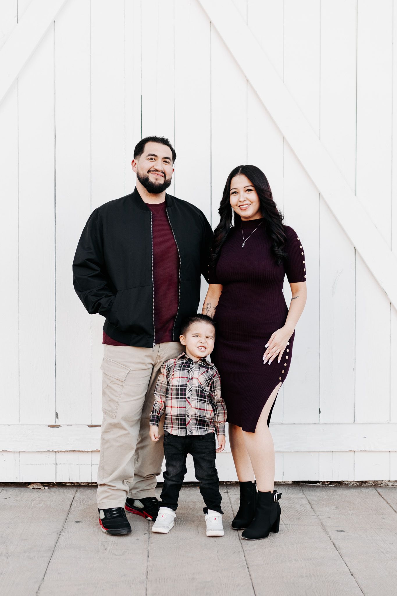 A family of three poses together against a white wall wearing coordinated burgundy and black outfits.