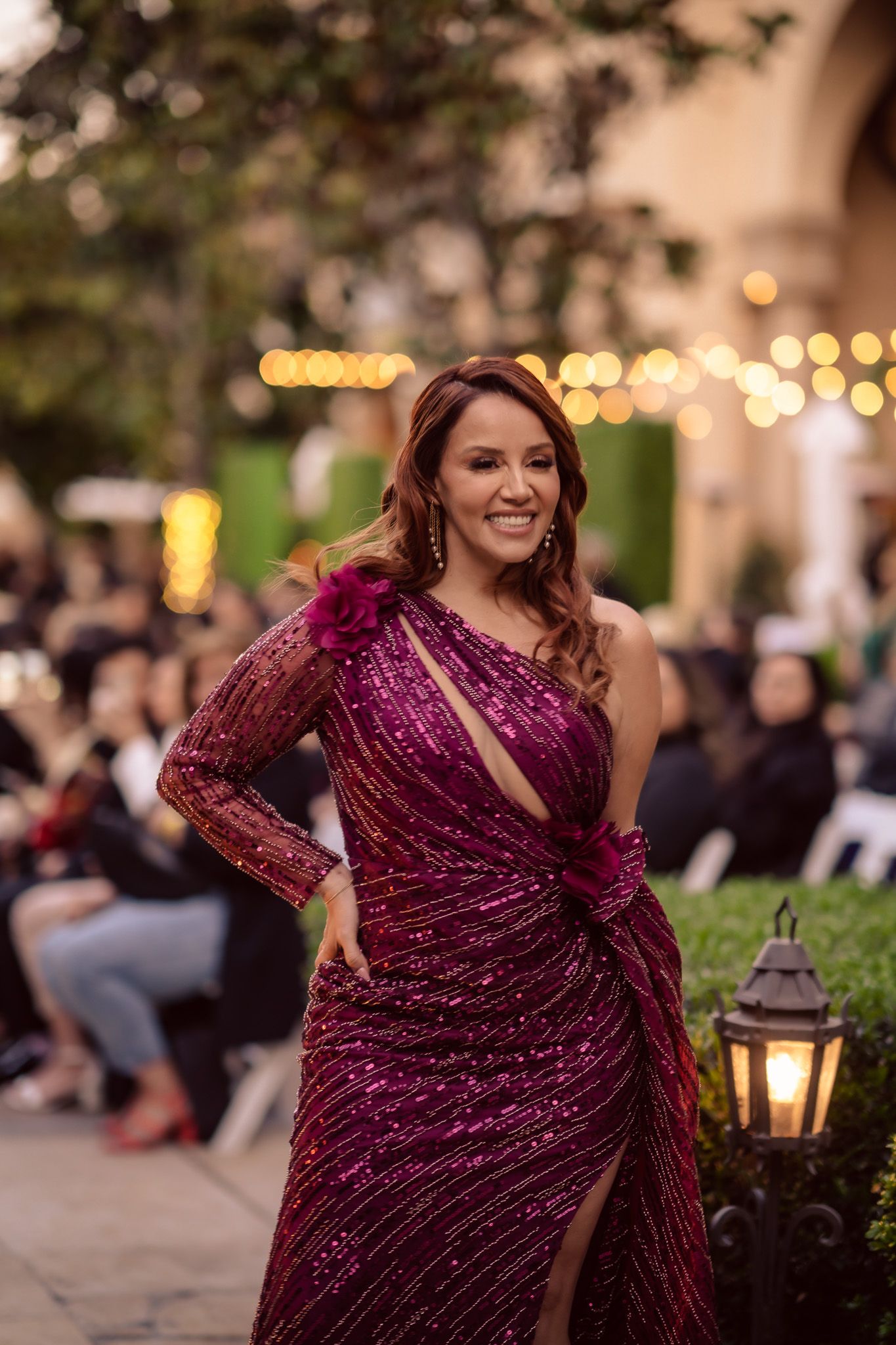 A woman in a sparkling burgundy evening gown poses at an outdoor event with string lights and lanterns in the background.