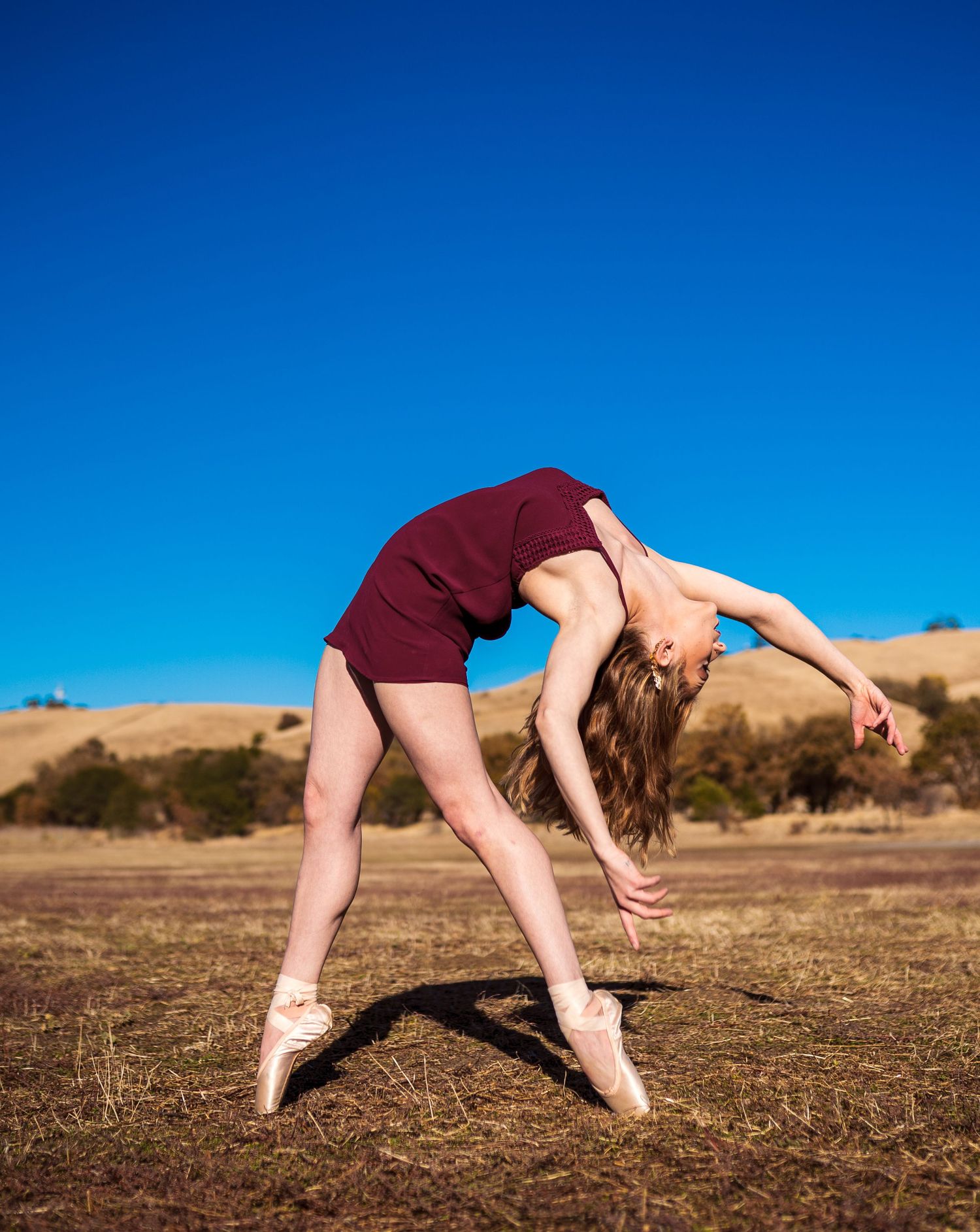 Dancer in burgundy leotard performs graceful backbend on pointe shoes against bright blue sky in outdoor setting.