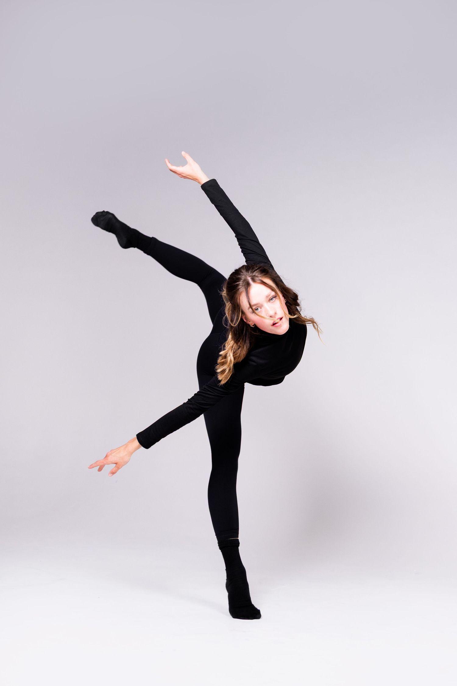 A dancer in a black leotard performs an elegant arabesque pose against a light gray studio background.