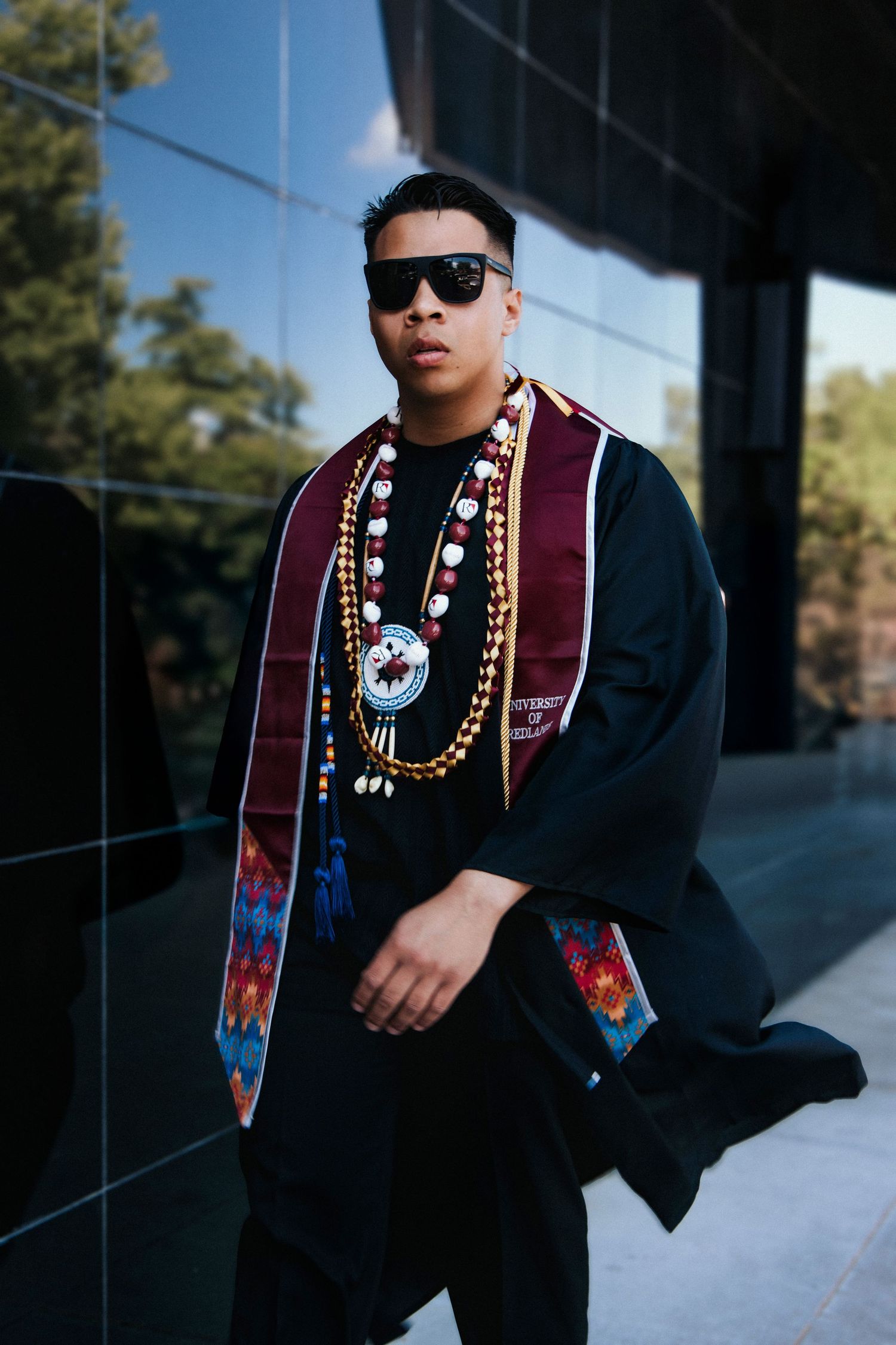 A person in graduation attire wearing sunglasses and traditional Native American beaded necklaces stands outside a modern building.