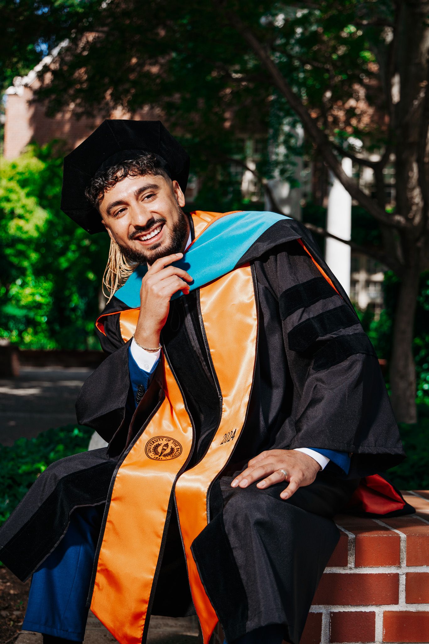 A graduate in academic regalia with orange and black graduation robes sits smiling on a brick wall outdoors.