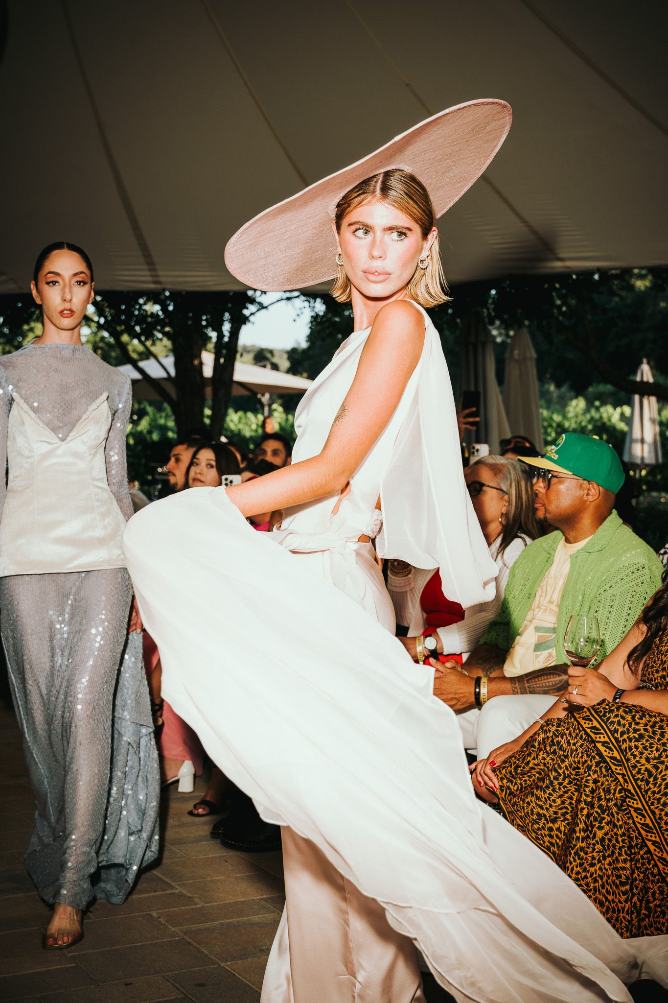 Models showcase elegant evening wear at a fashion show with one wearing a flowing white gown and dramatic wide-brim hat.