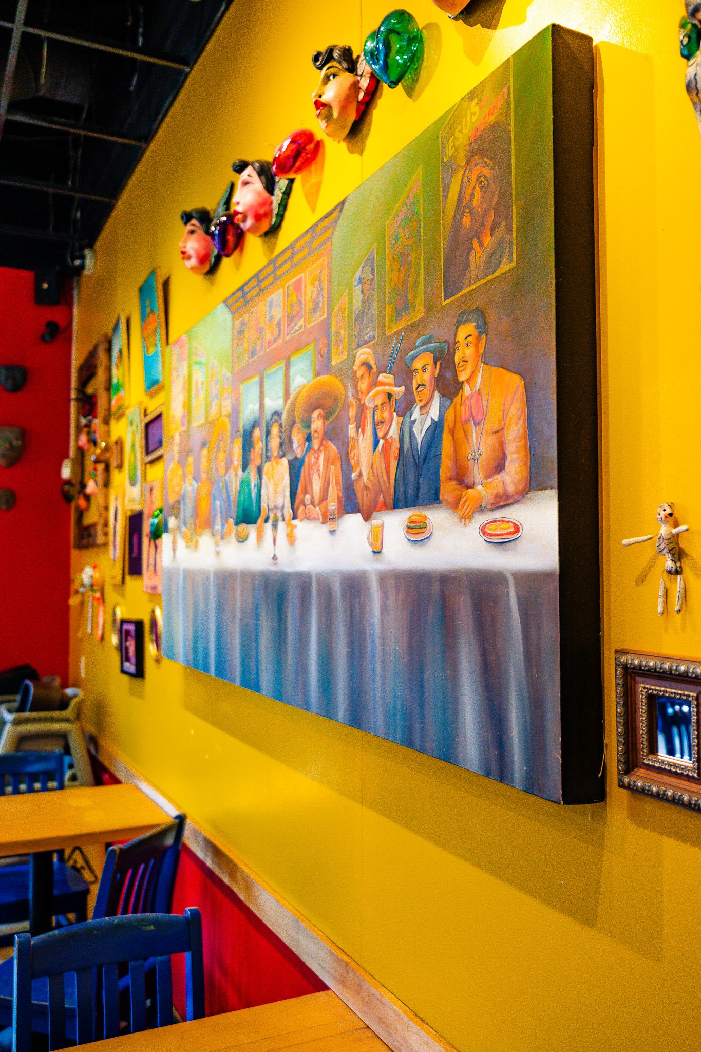 Colorful restaurant interior with yellow walls features pop art decor and a large mural depicting diners at a long table.