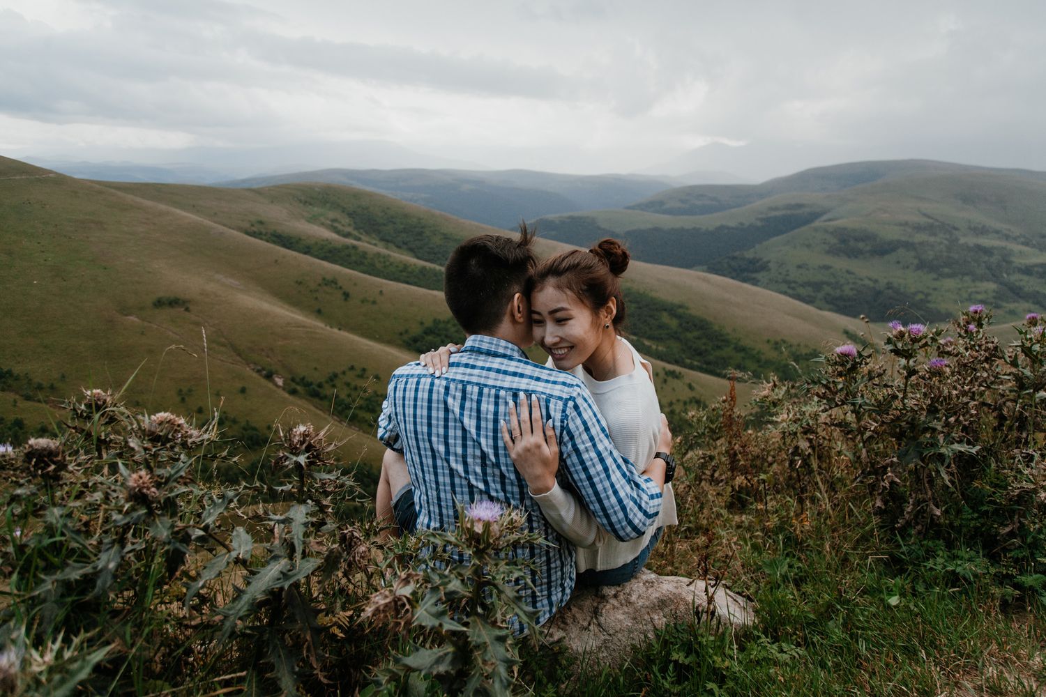 A couple embracing while sitting on rocks overlooking rolling hills.