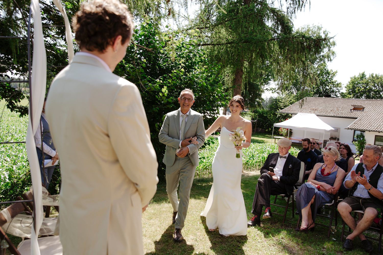 Garden wedding ceremony with guests gathered under trees and umbrellas.