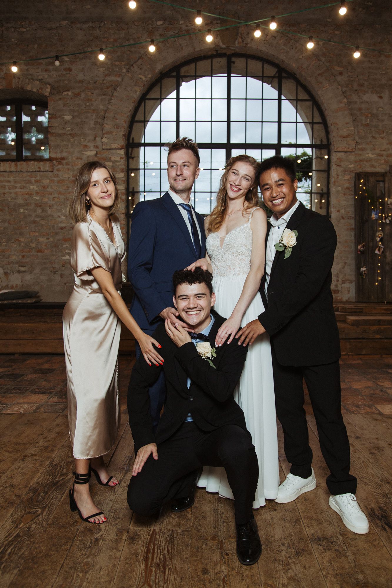 A joyful wedding party posing in front of an arched window in an industrial brick venue with string lights.