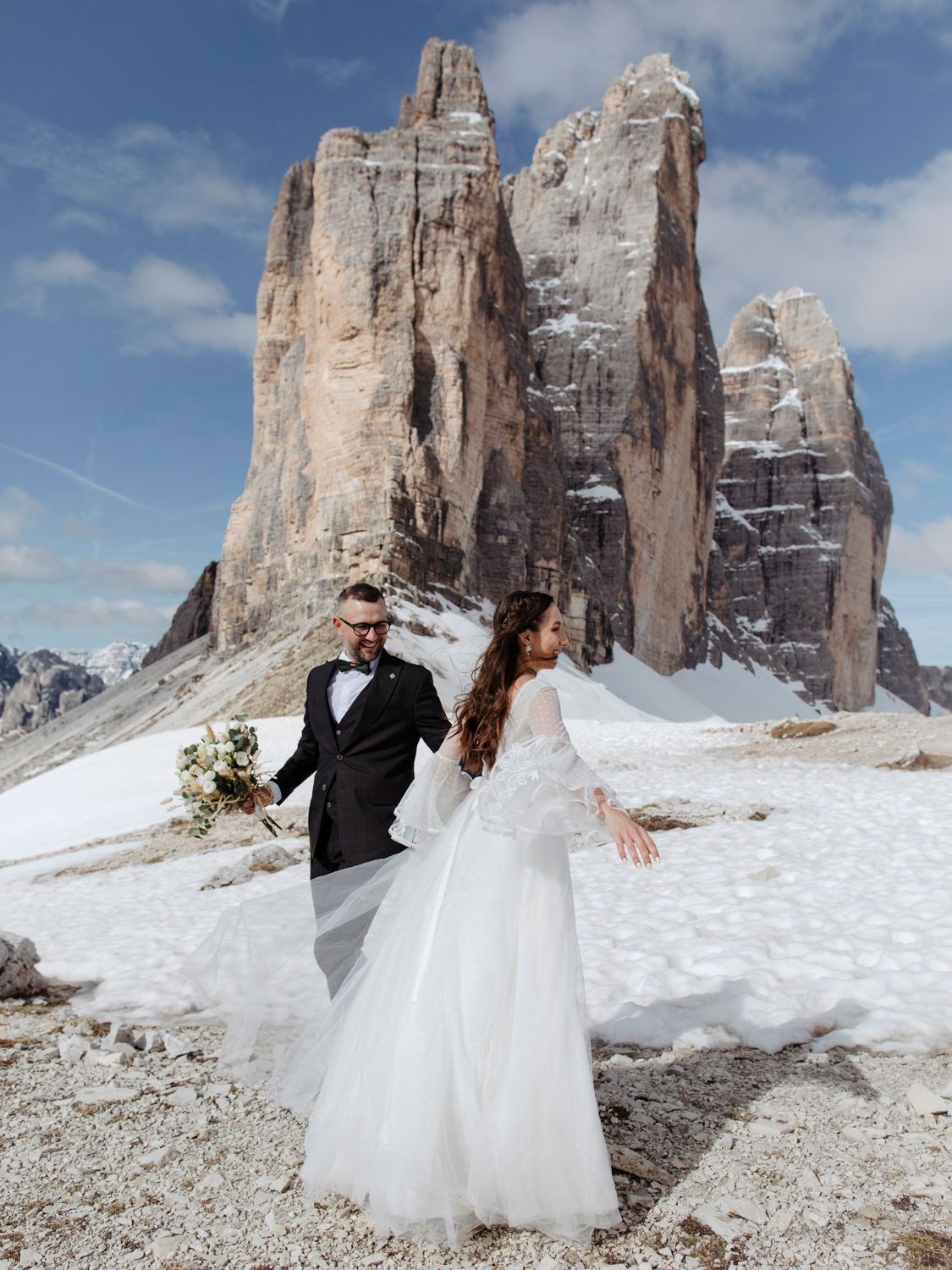 Wedding couple posing in formal attire against snowy Dolomites mountain backdrop.