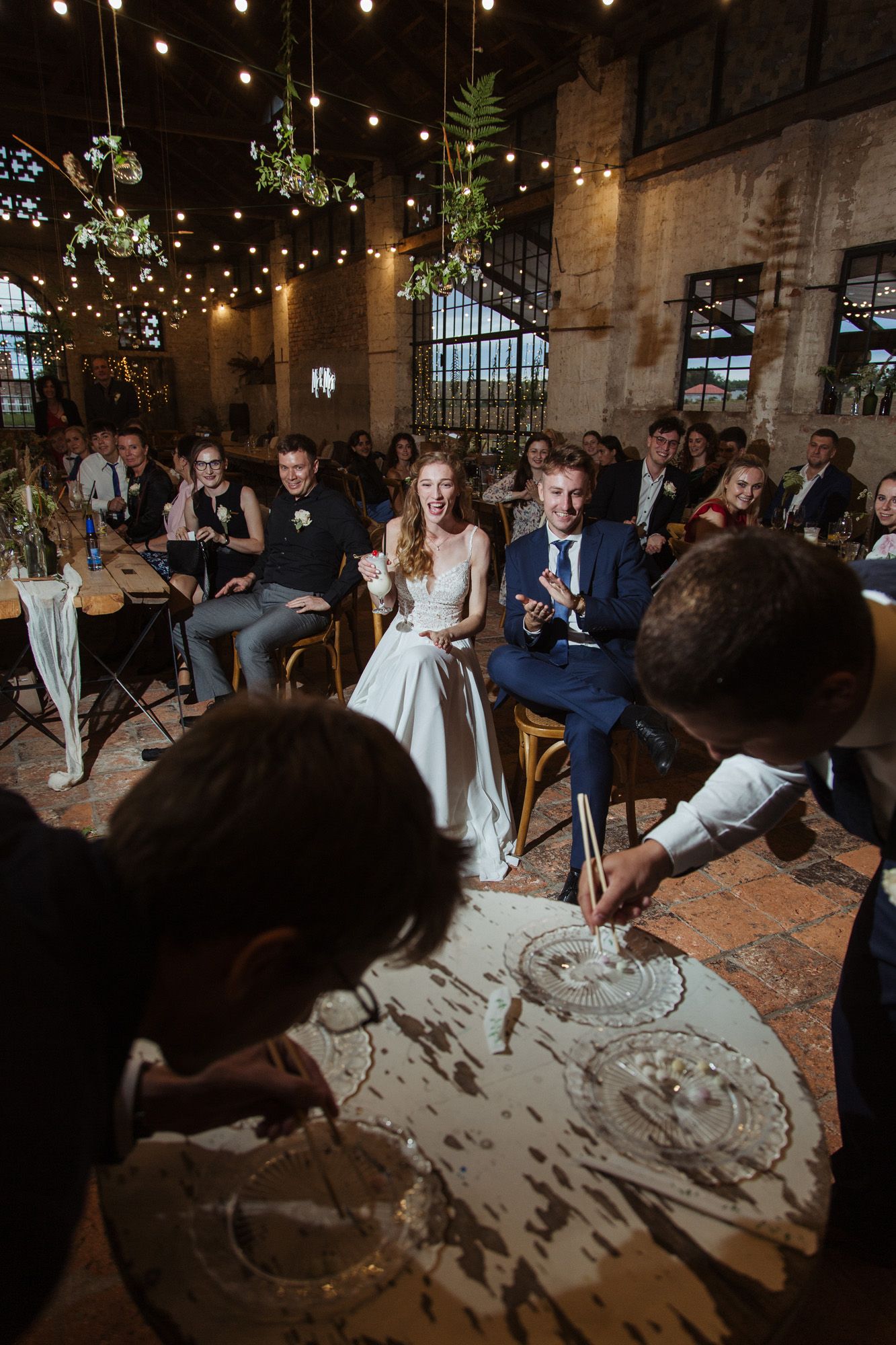 Wedding reception in an industrial brick venue with string lights and hanging greenery.
