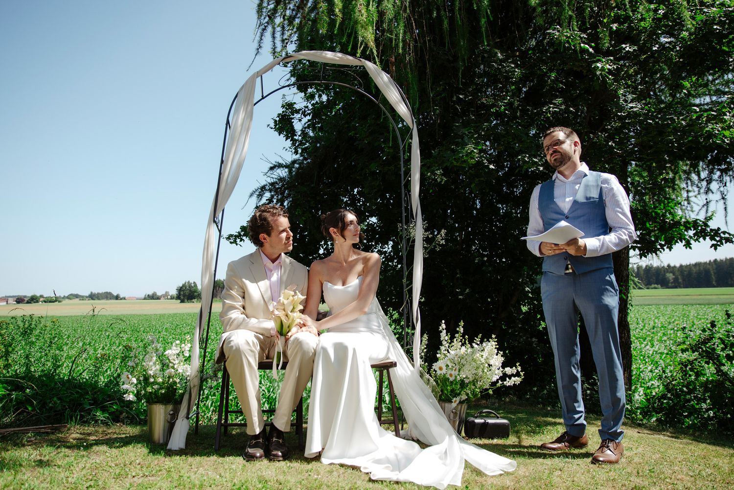 Outdoor wedding ceremony under a willow tree with guests seated on white chairs.