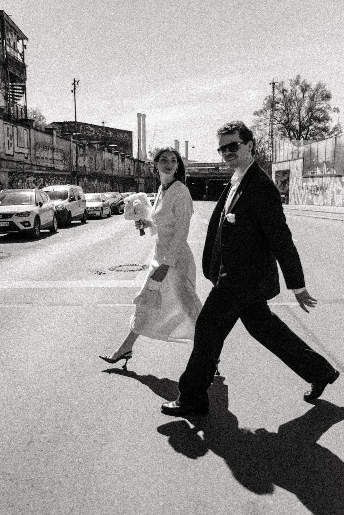 A candid street photo of a couple walking confidently in wedding attire with urban backdrop.