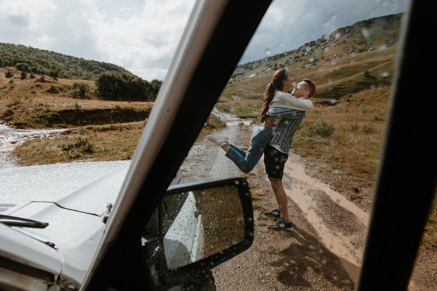 Car mirror reflection shows a playful lift pose on a mountain road.