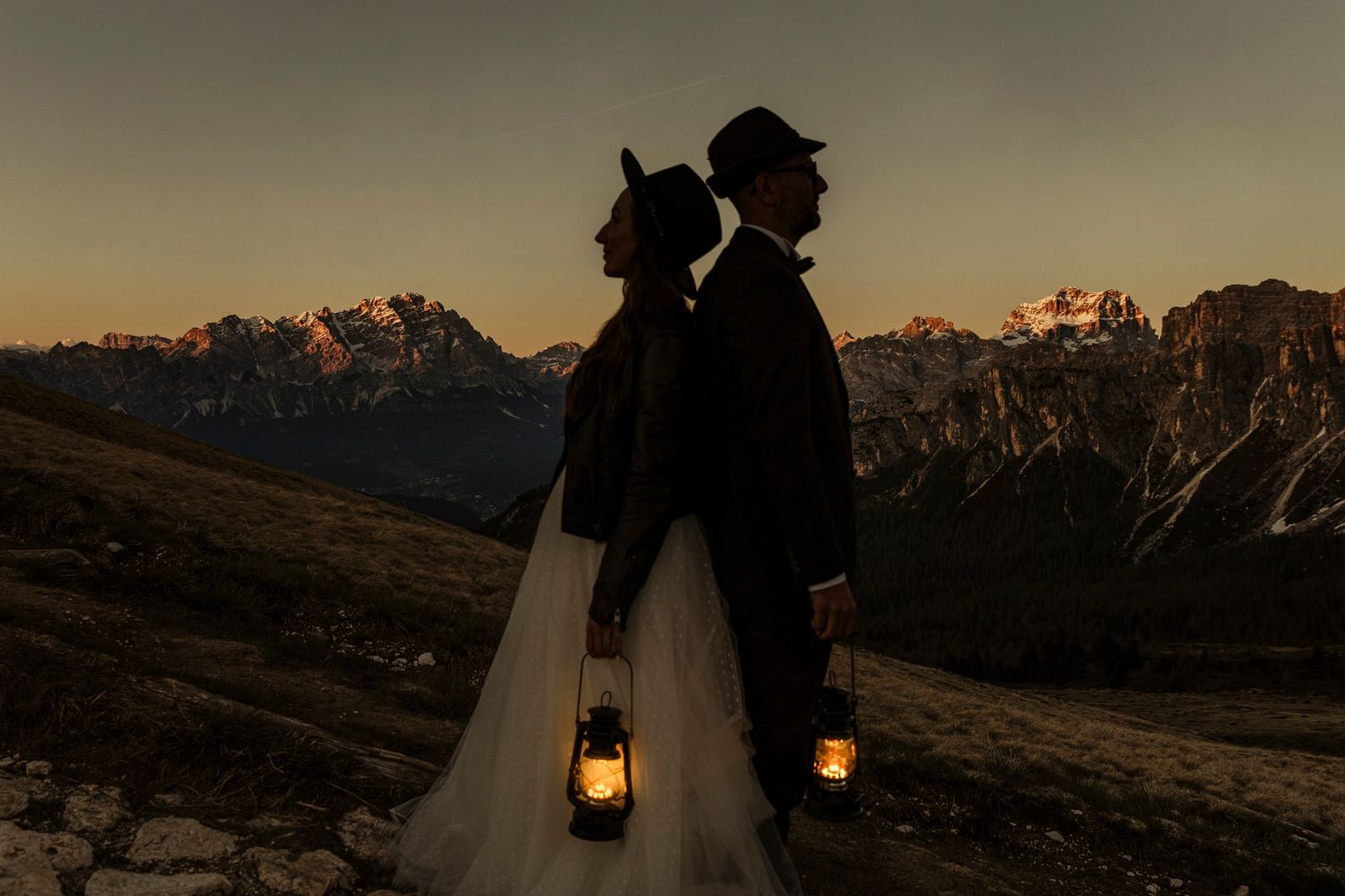 Silhouettes of two figures holding lanterns against mountain backdrop at dusk.