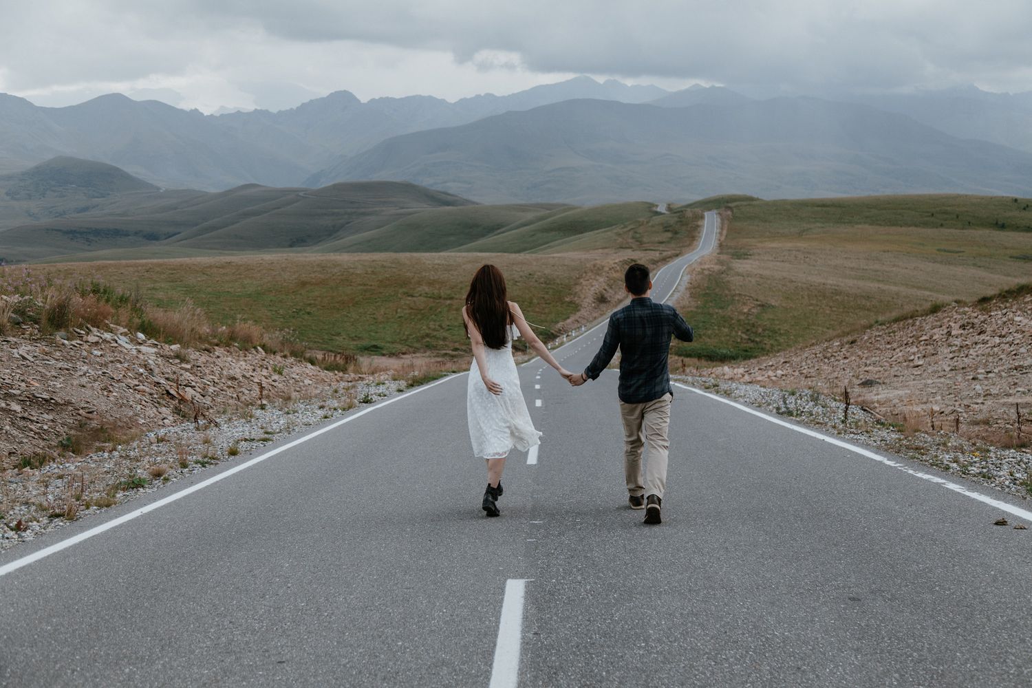 A couple runs hand in hand down an empty mountain road surrounded by rolling hills and cloudy skies.